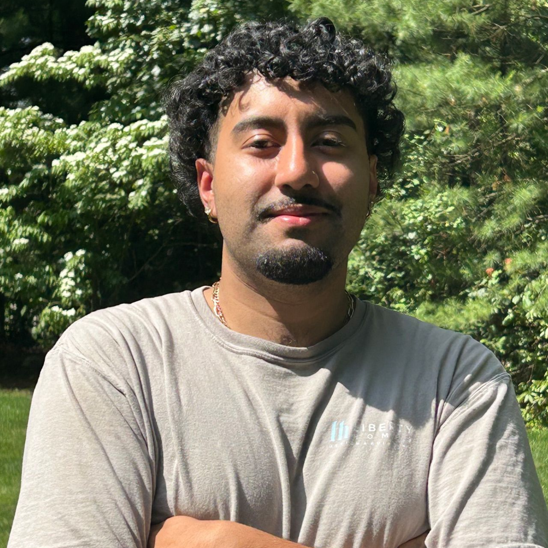 Person with curly hair and a gray shirt standing outdoors in sunlight with green trees behind.