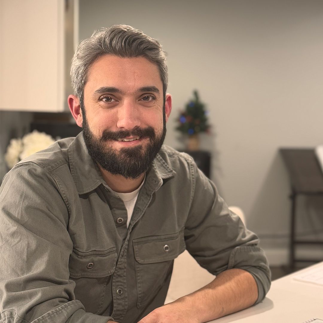 Smiling bearded man in a gray shirt sitting at a table in a softly lit room