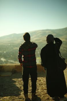 Dos personas de pie en una colina, contemplando un valle de montaña iluminado por el sol al amanecer.