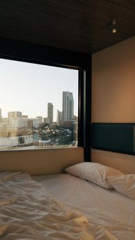 Habitación con cama sin hacer junto a una gran ventana con vistas al horizonte de la ciudad al atardecer.