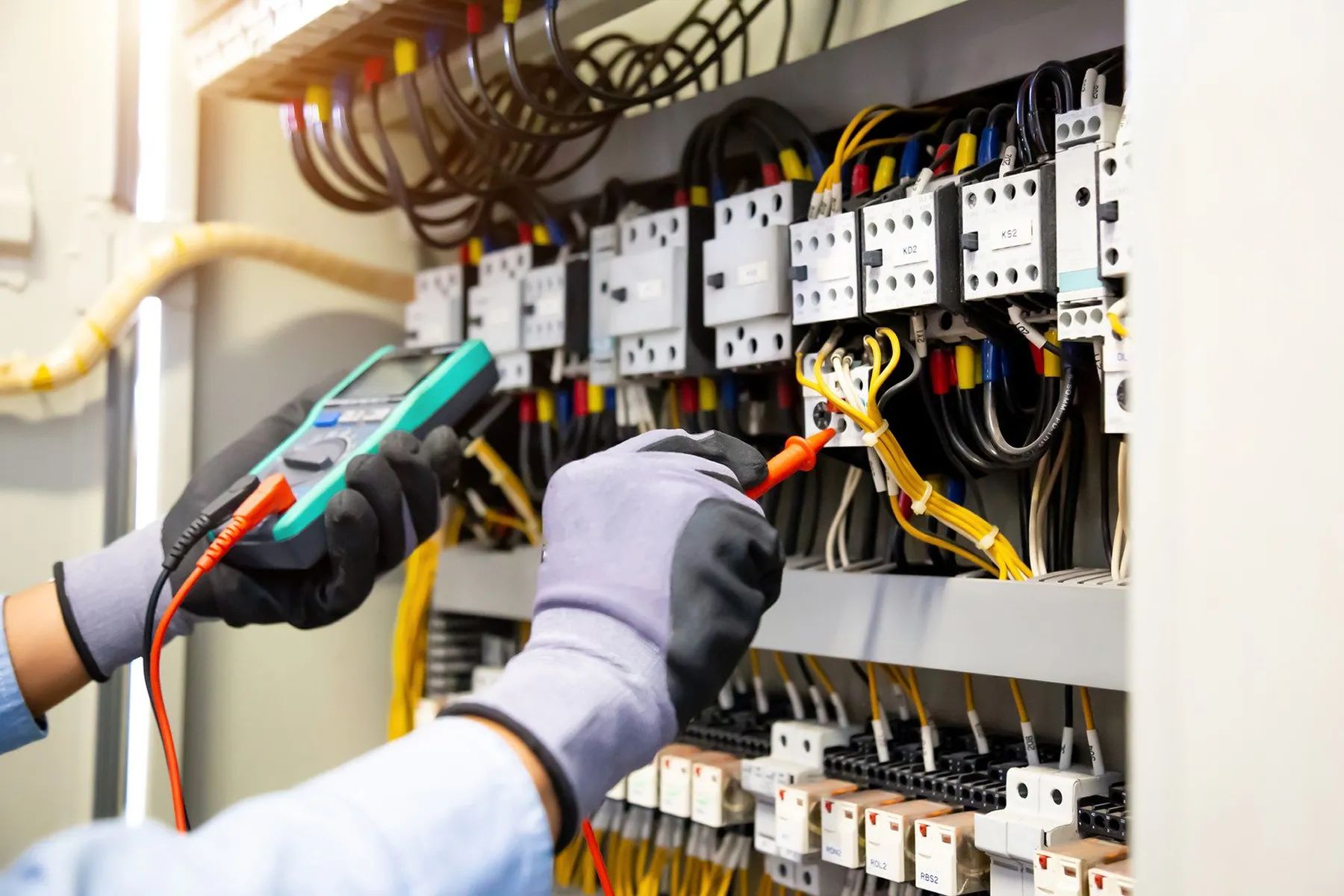 Electrician using a multimeter to inspect an electrical panel with wires and circuit breakers.