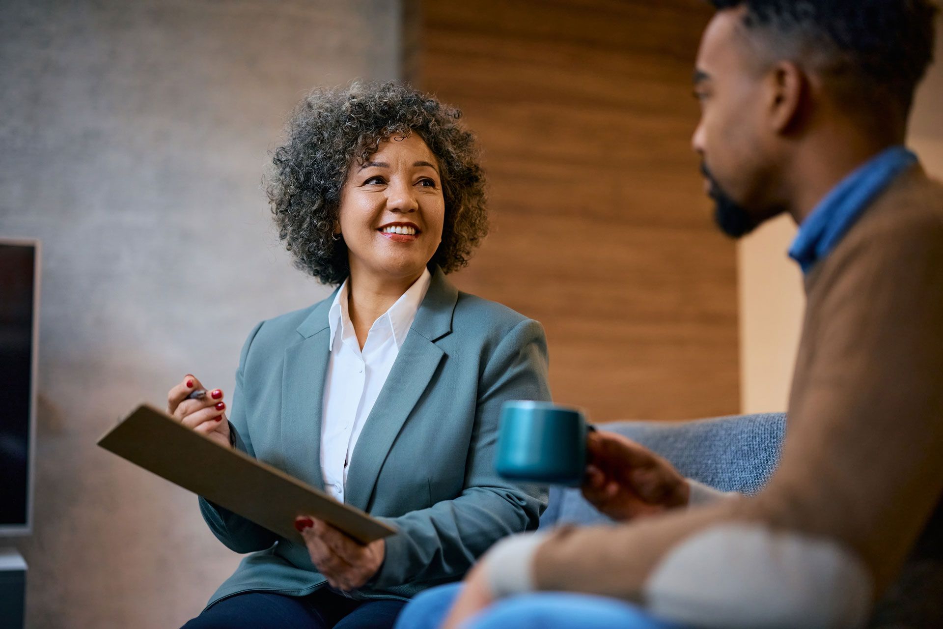 A professional in a blazer holds a clipboard and smiles at a seated person holding a blue mug during a consultation.