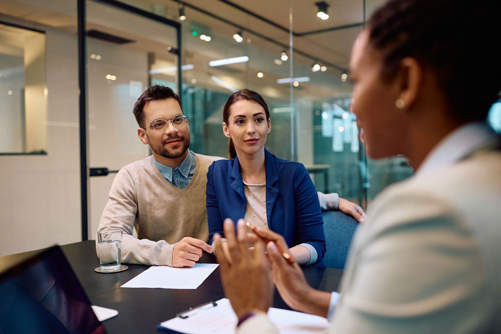 A person leads a professional meeting with two attentive colleagues in a modern, glass-walled office boardroom.