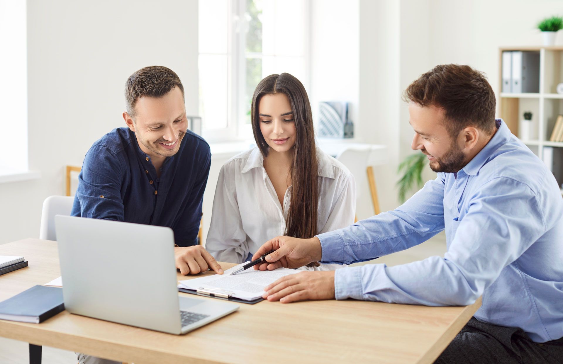 Three professionals in a modern office meeting, reviewing documents on a desk with a laptop.