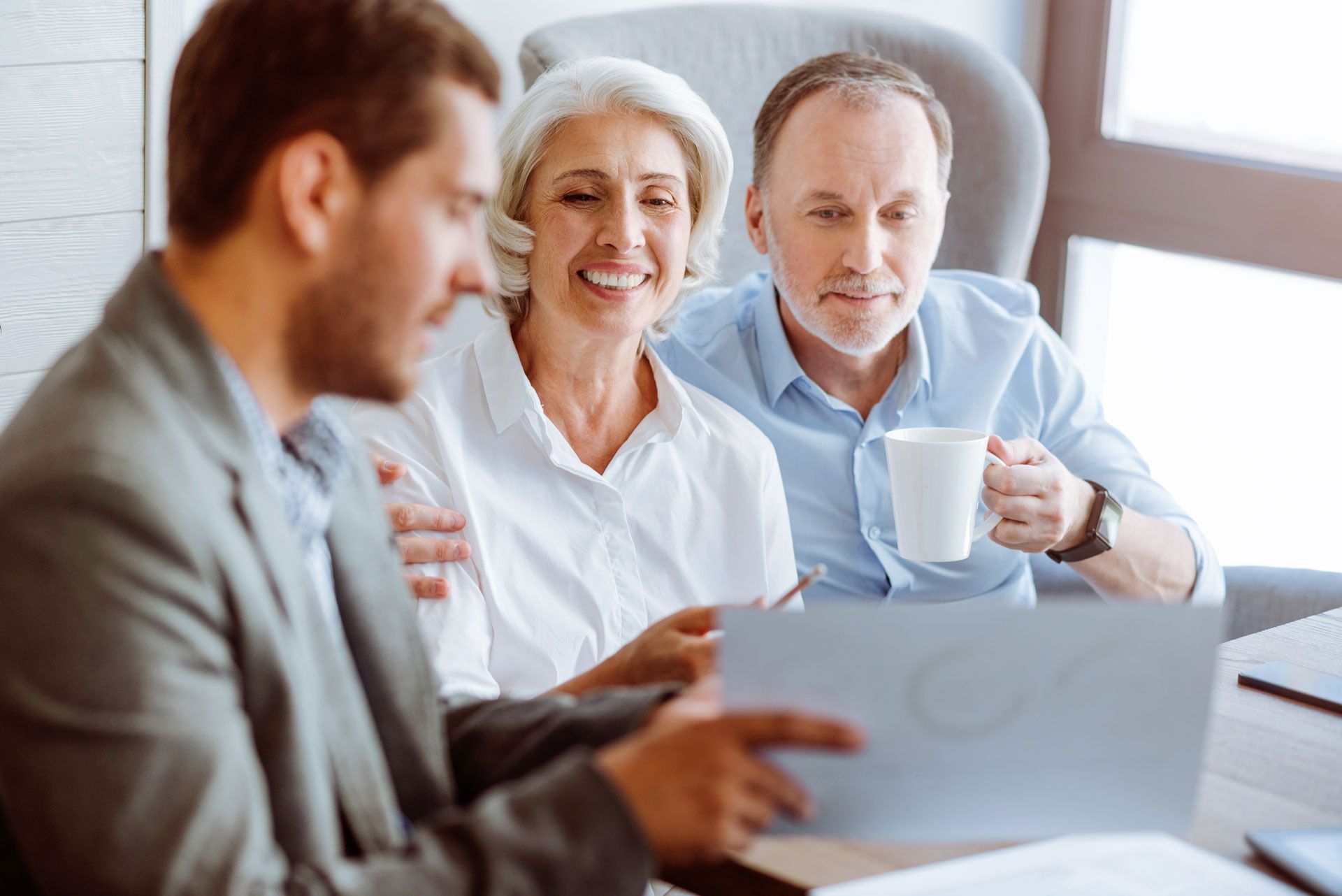 A financial advisor reviews documents with a smiling couple holding a coffee mug in a well-lit office.