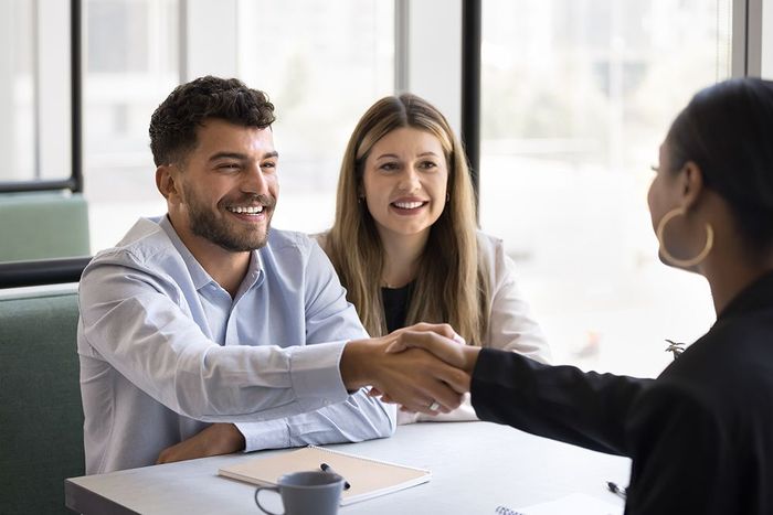 Man shaking hands with a woman, smiling; a woman watches from the background at a desk. Bright, neutral setting.