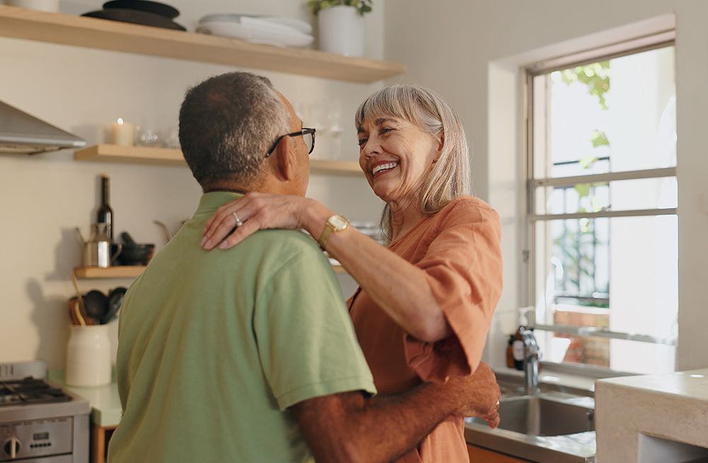 Older couple dancing in a kitchen, woman laughing, wearing orange dress, man in green shirt, light and airy setting.