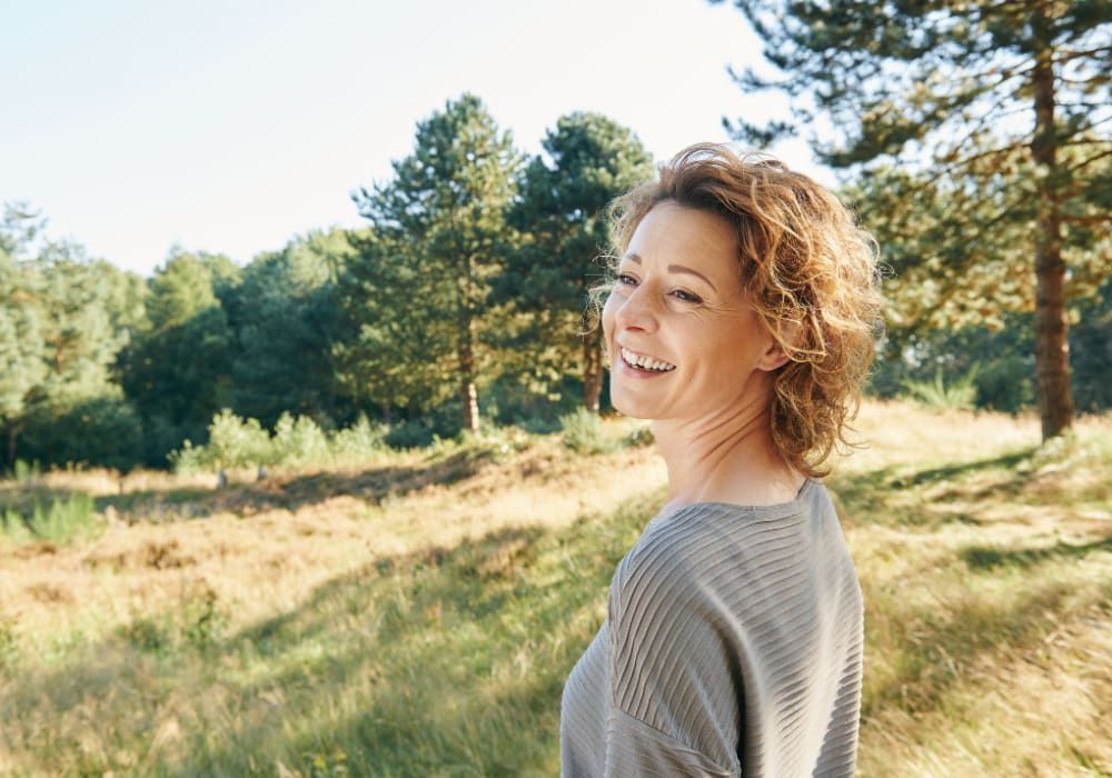 Woman with curly hair smiles, looking back over her shoulder, outdoors in a sunny field of grass and trees.