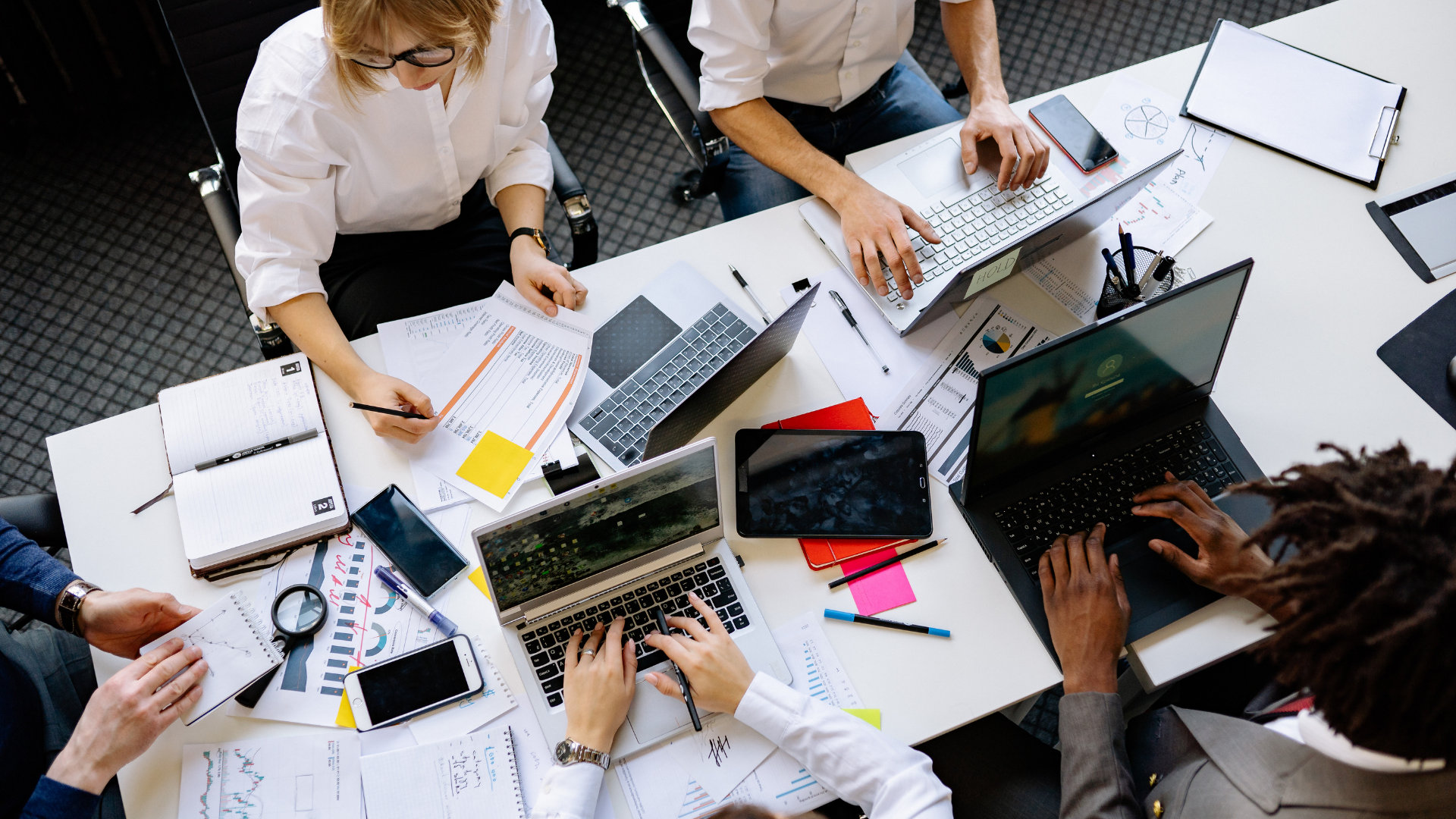 People collaborating around a table with laptops, papers, and charts in a bright office meeting.