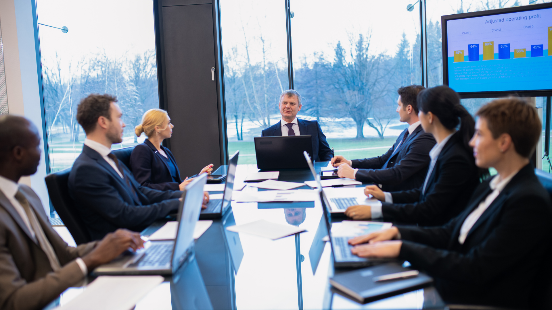 Business meeting in a glass conference room, with people seated around a table and a presenter at the front.