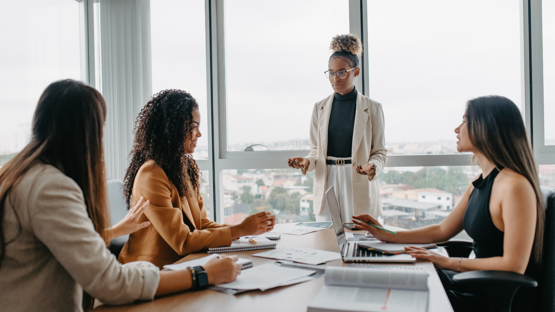 Business meeting in a bright conference room with three people seated and one person standing by the window.