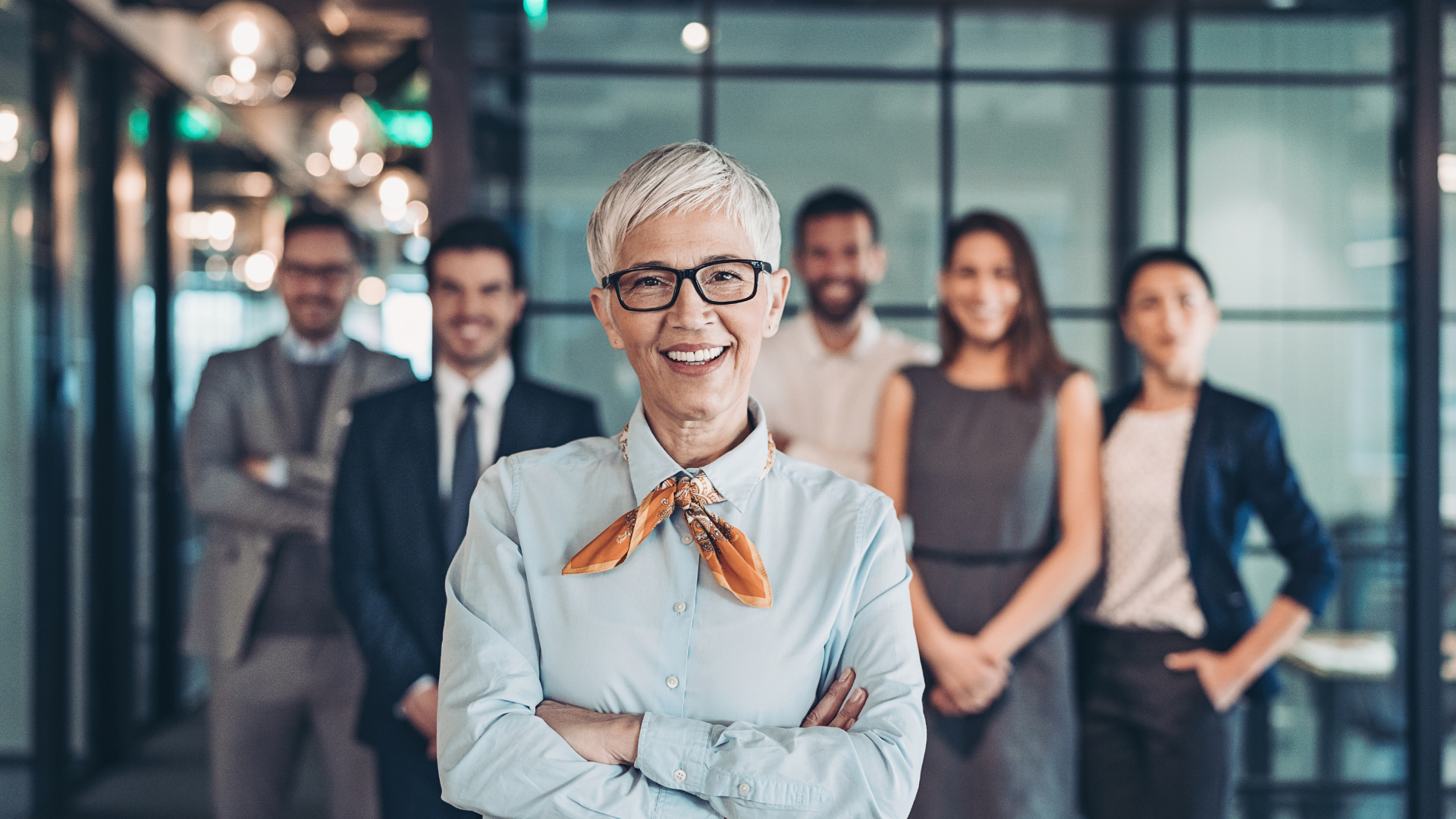 Smiling person with glasses and orange scarf stands in front of a group in an office setting