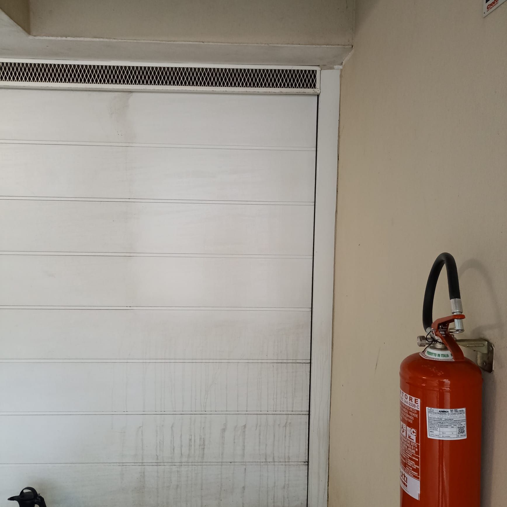 Un estintore rosso montato su una parete beige accanto a una porta bianca con pannelli orizzontali e una griglia di ventilazione nera.