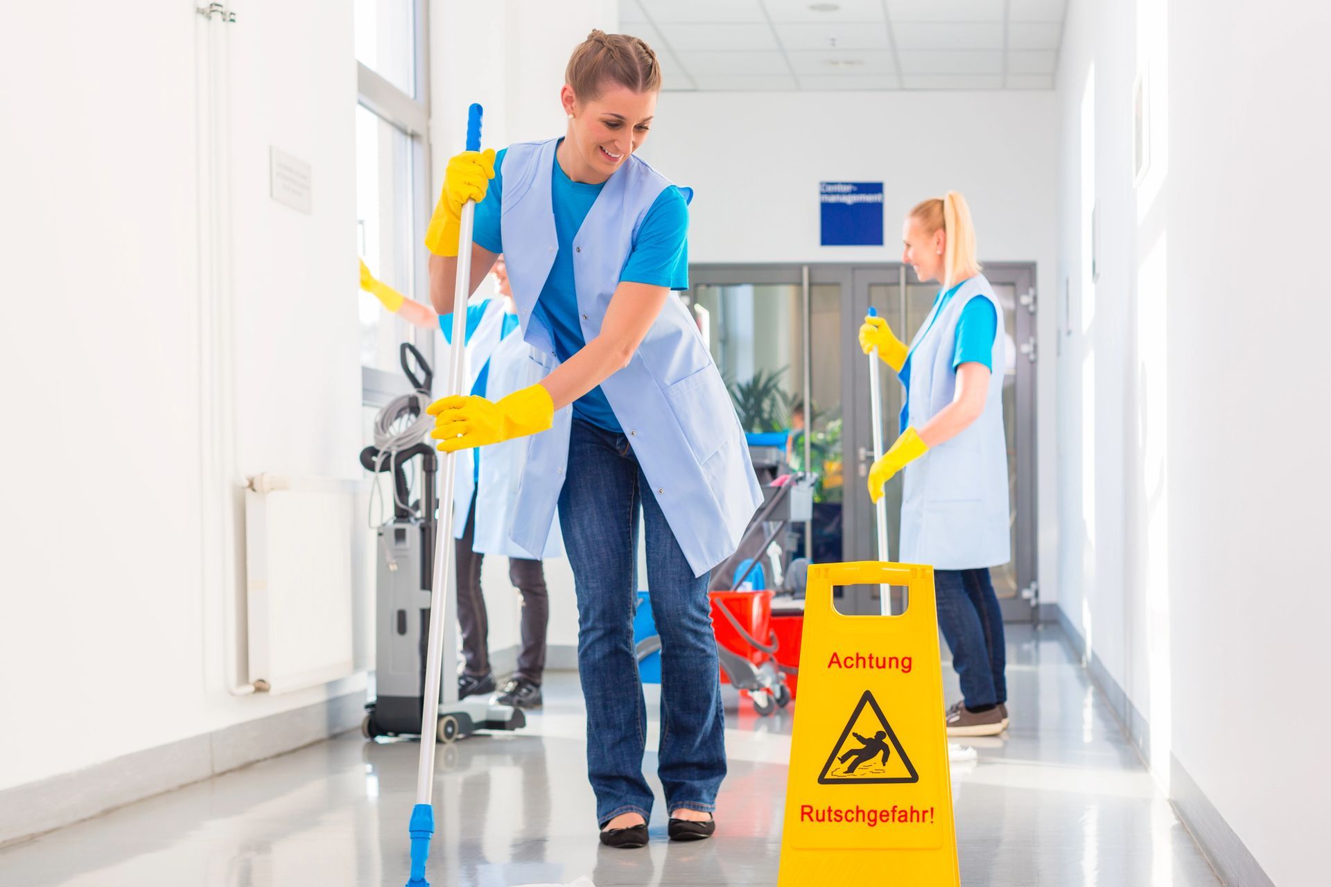 Tre addetti alle pulizie in uniforme blu lavano il pavimento di un corridoio pulito e luminoso, con un cartello giallo di avvertimento in primo piano.