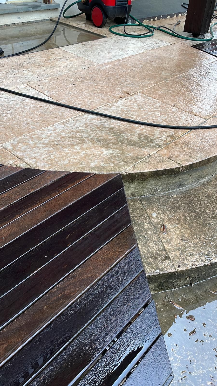 Una ripresa dall'alto mostra un patio in cemento bagnato e ruvido in fase di pulizia, con una superficie in legno scuro a listelli in primo piano.