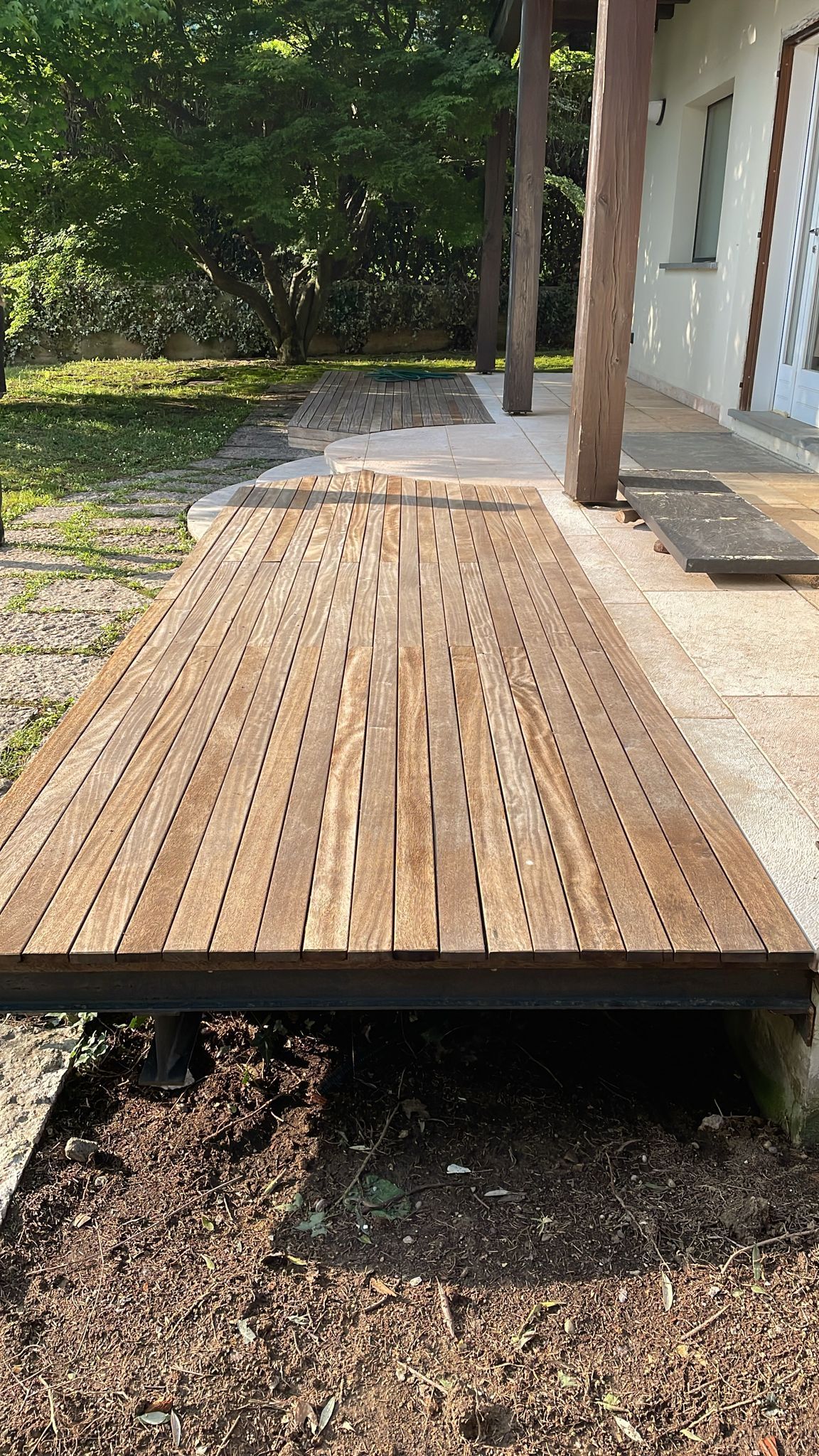 Una terrazza in legno con doghe e fori, rialzata rispetto a un terreno ghiaioso, accanto a una casa con pali di legno e un muro bianco.