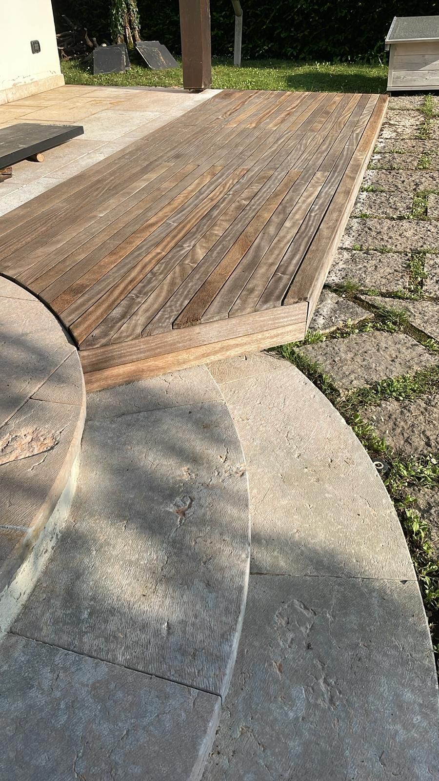 Una piattaforma in legno si trova su un patio in pietra con gradini curvi, delimitato da un sentiero di ghiaia e pietra in un cortile.