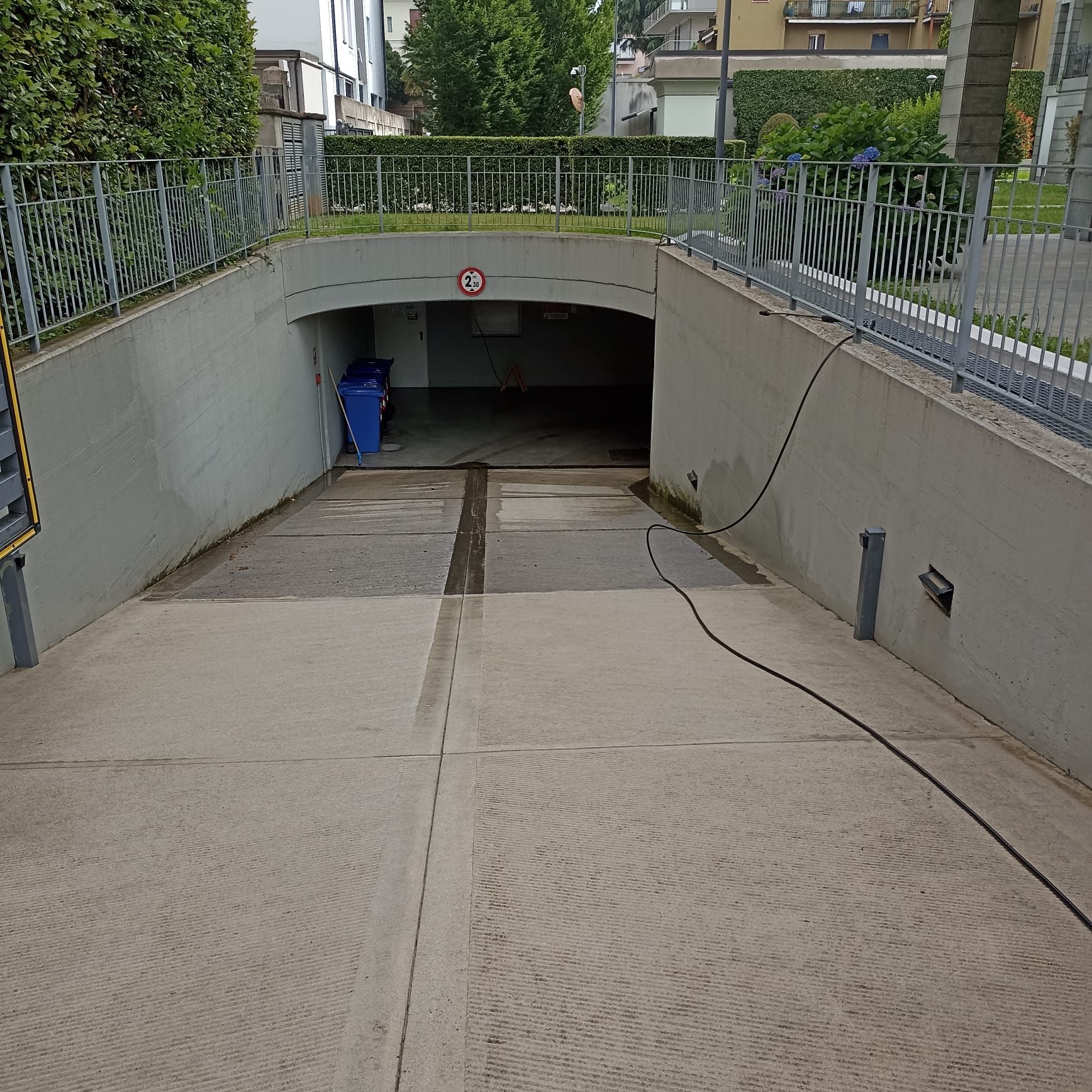 Un vialetto in cemento in pendenza conduce all'ingresso di un garage sotterraneo buio, con un piccolo bidone blu per la raccolta differenziata.