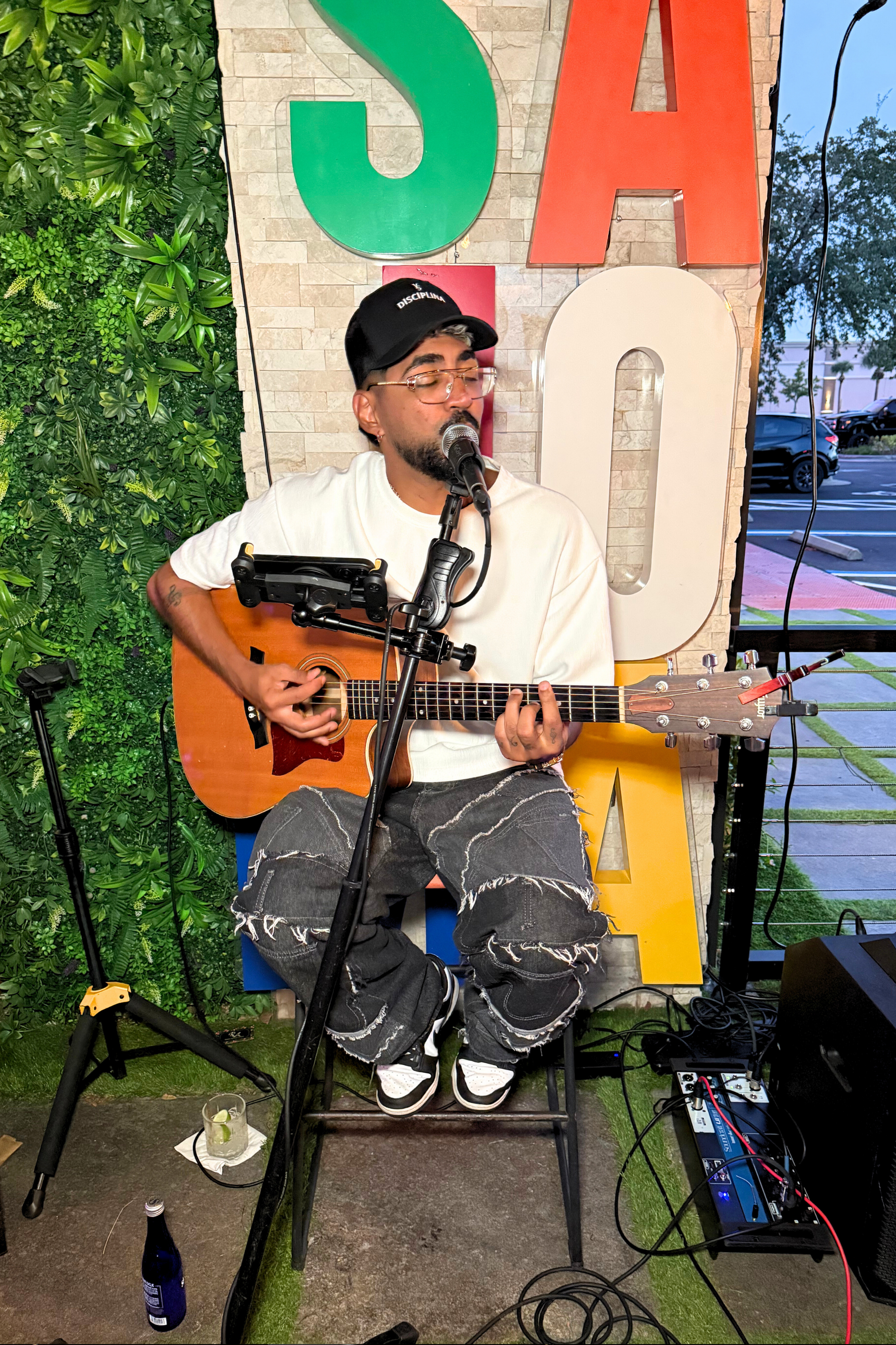 Man playing acoustic guitar, wearing a cap and tie-dye pants, in front of a sign outdoors.