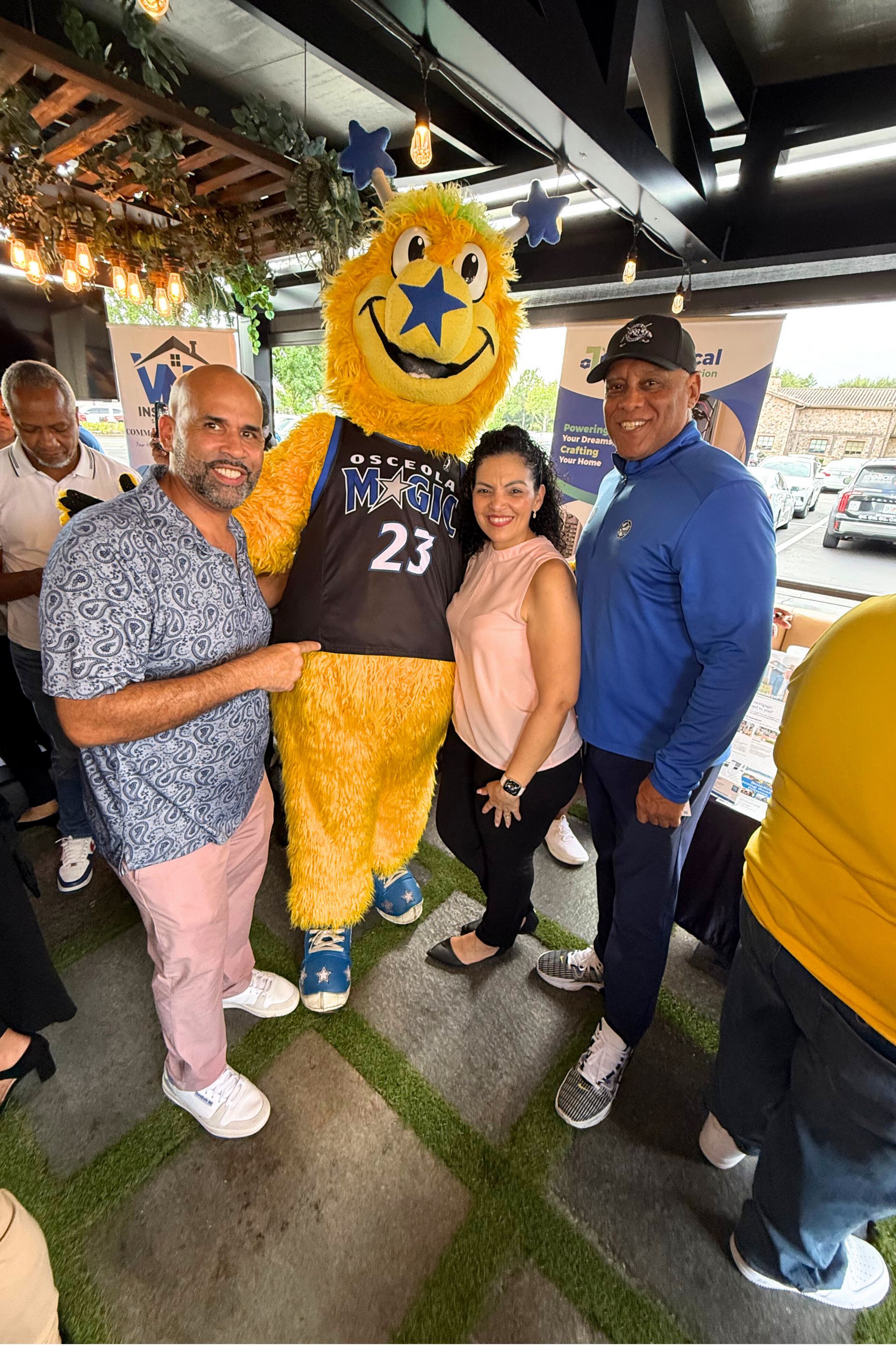 People pose with a mascot at an outdoor event. Man in pink pants, woman, and man in blue smile. Mascot is yellow.