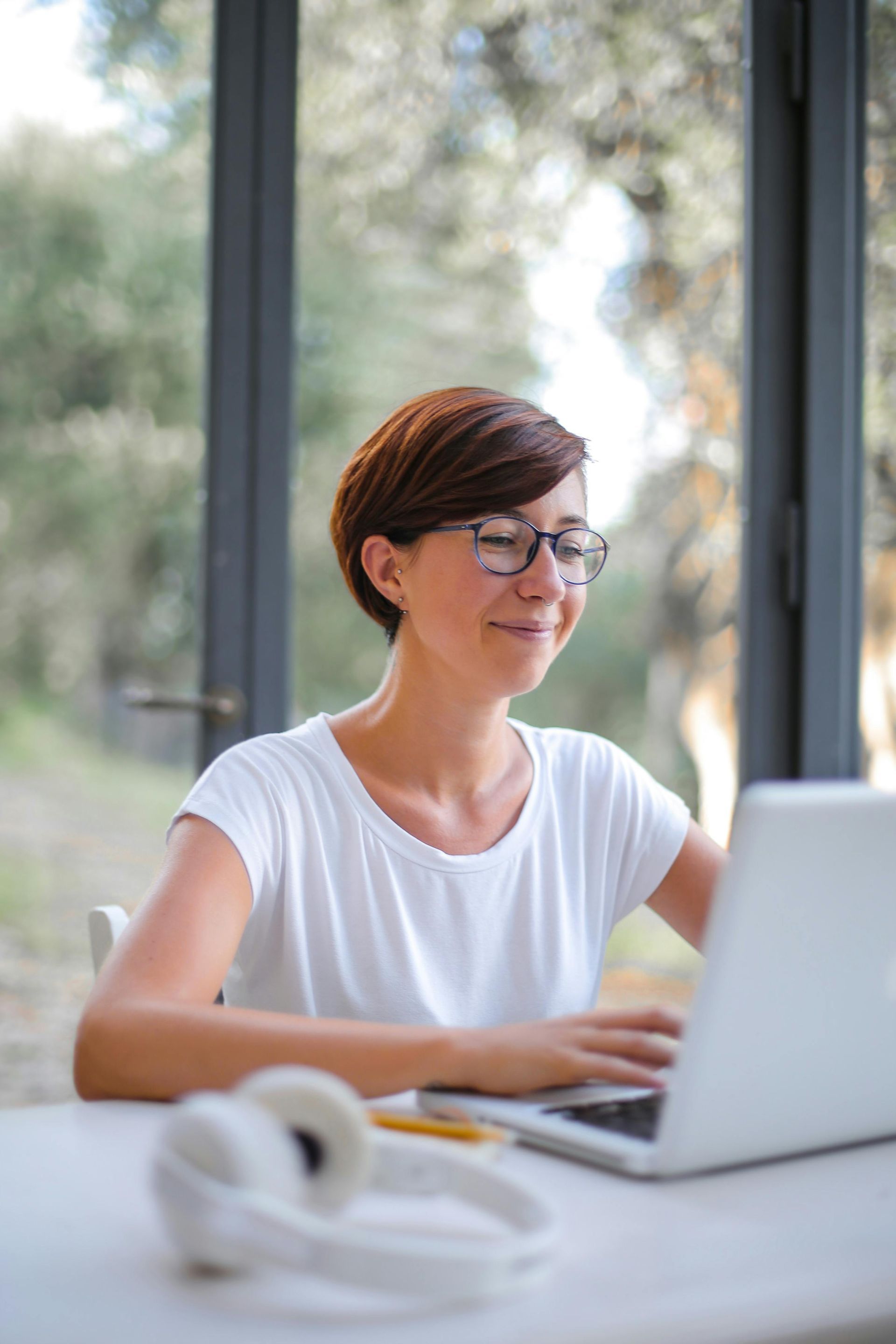 Woman with glasses smiles while typing on a laptop at a white table, headphones nearby.