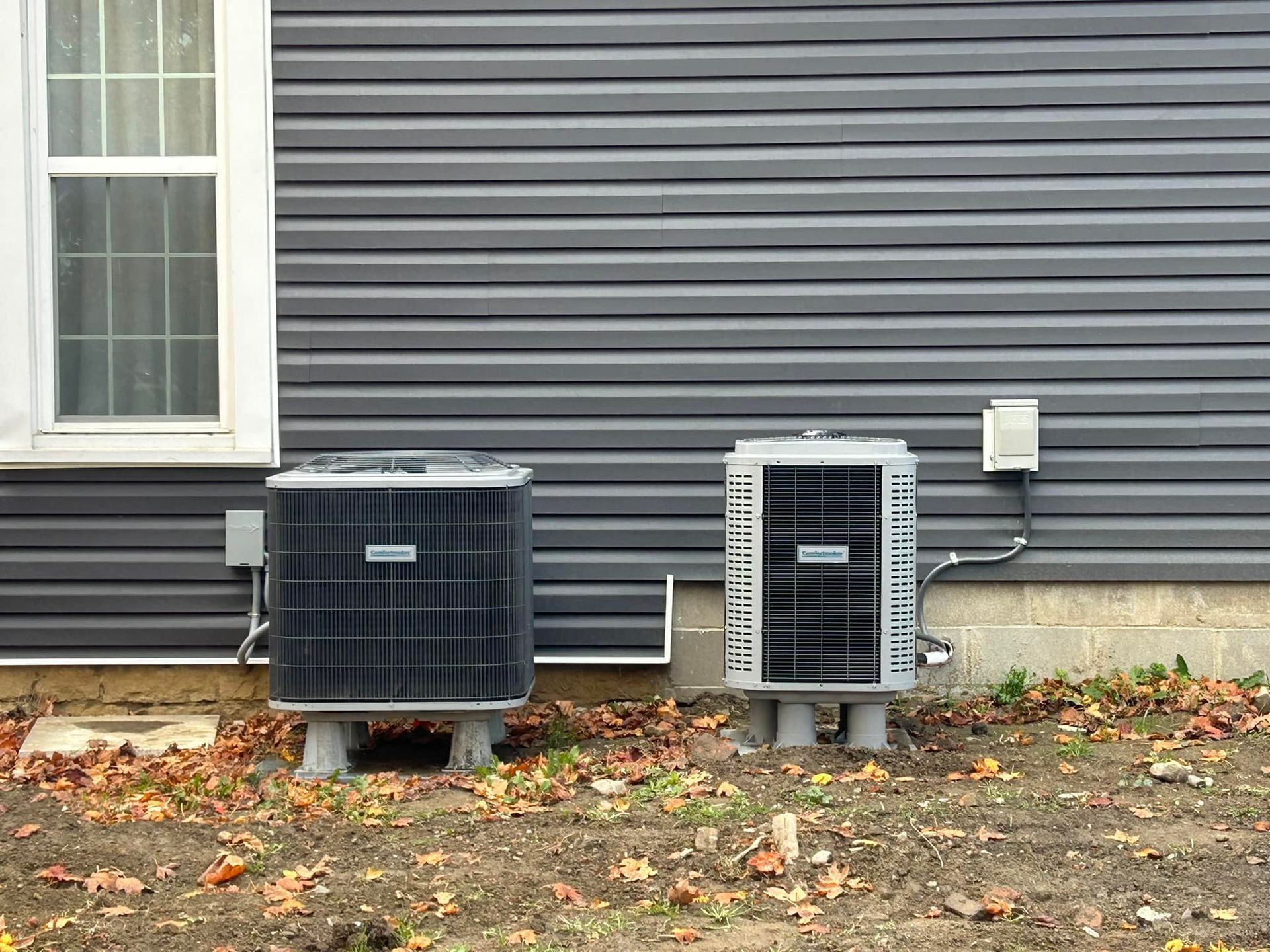 Two HVAC units sit on concrete pads against the gray siding of a house with a window on the left.