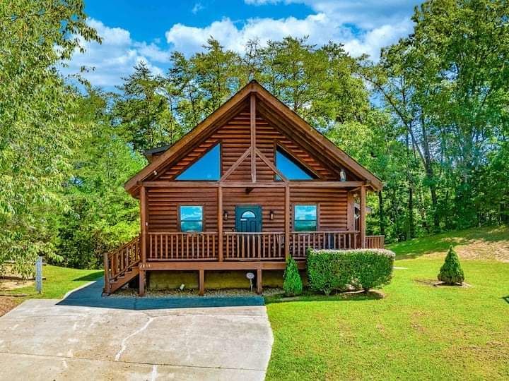 A brown wooden cabin sits on a grassy lawn surrounded by green trees under a blue sky with white clouds.