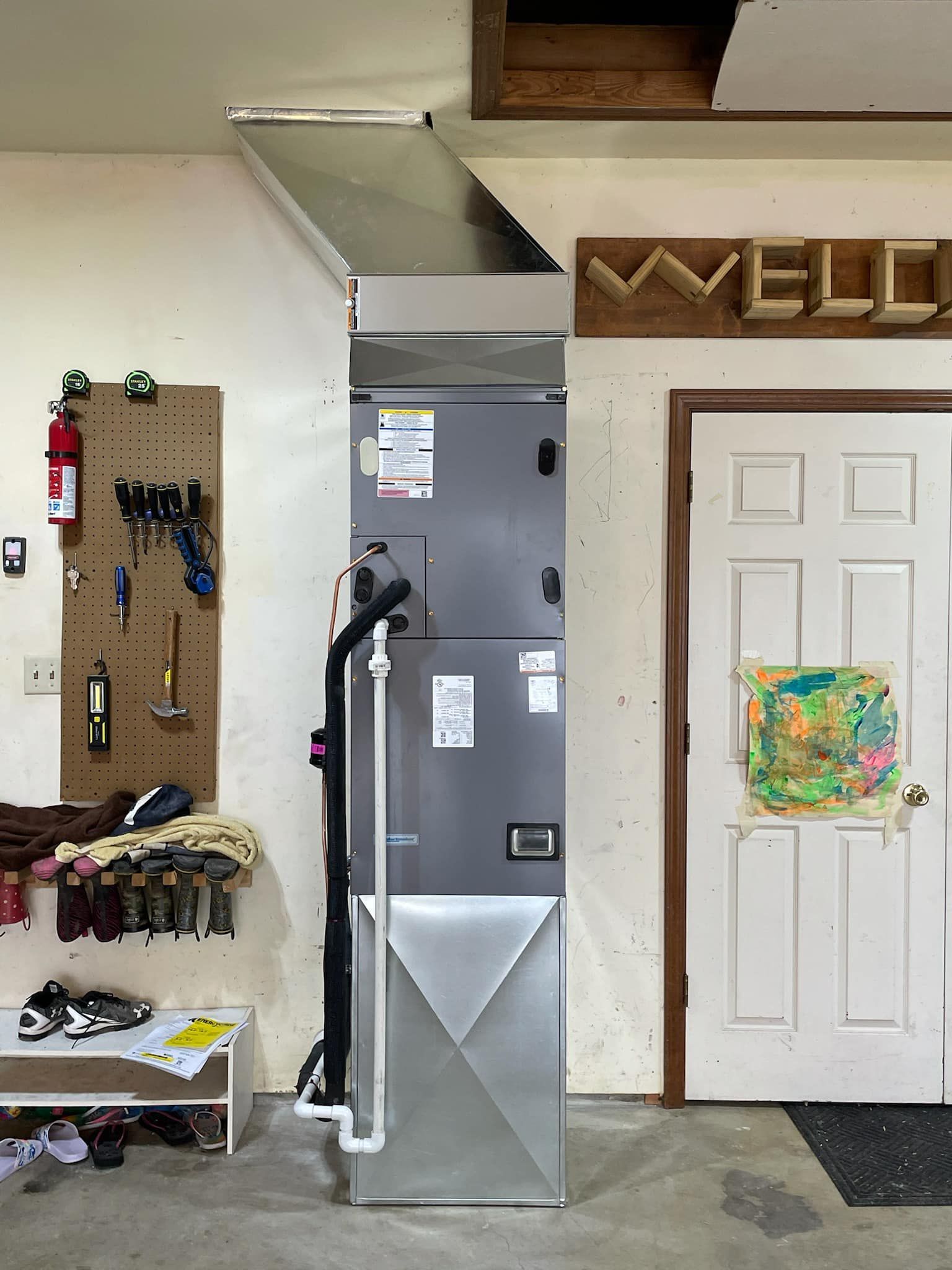 A gray furnace unit with metal ductwork stands against a white garage wall, next to a door and a pegboard tool organizer.