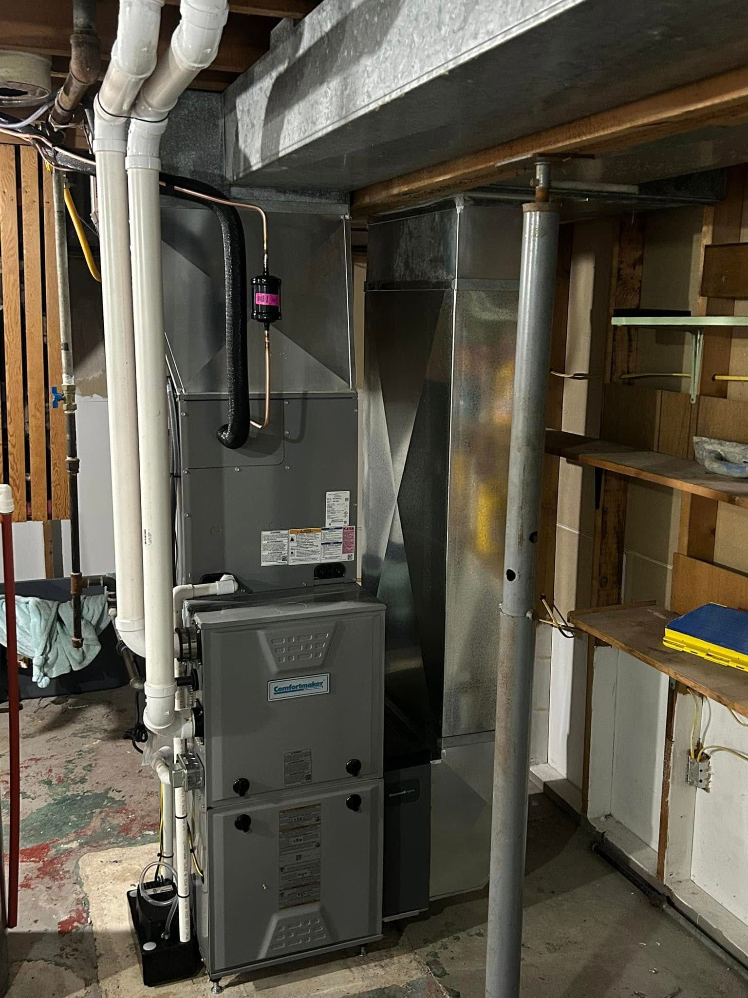 A gray HVAC furnace unit with white PVC vent pipes stands in a basement near a metal support pole and wooden shelves.