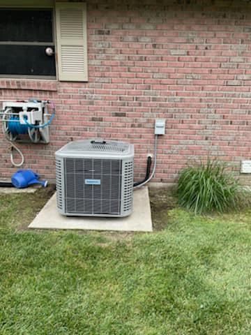 An outdoor air conditioning unit sits on a concrete pad next to a brick house wall near a water hose reel and tall grass.