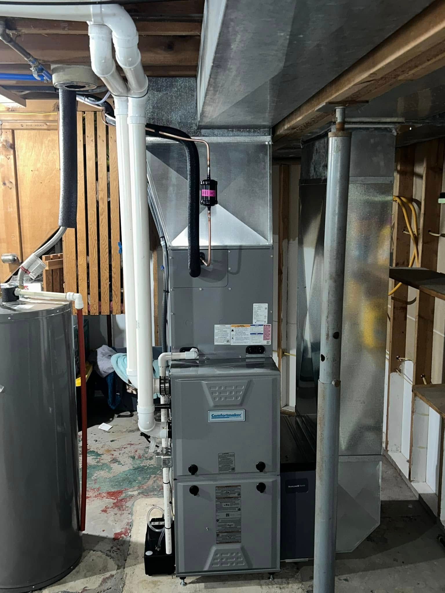 A gray high-efficiency furnace stands in a basement with white PVC intake and exhaust pipes and metal ductwork above.
