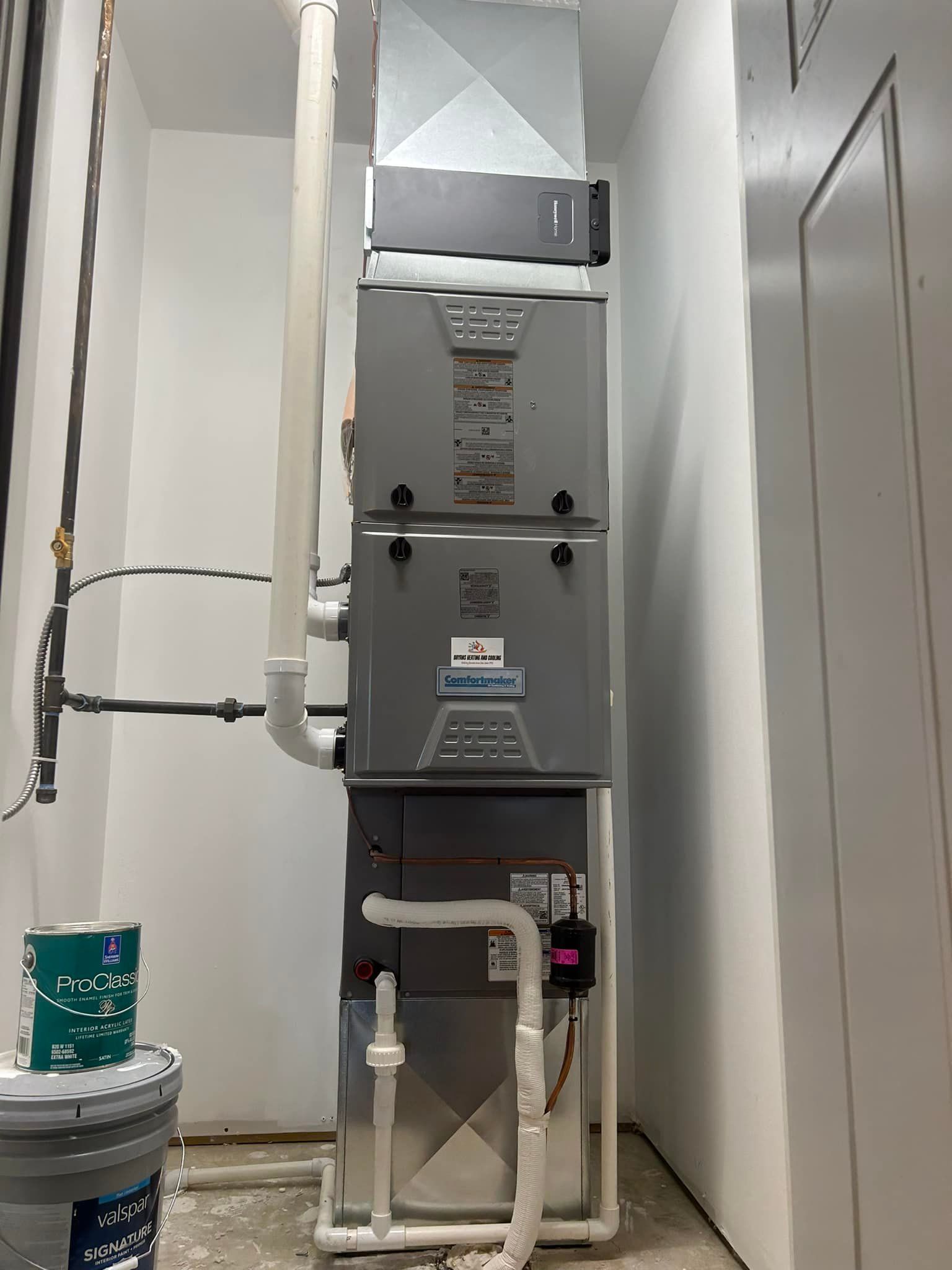 A vertical HVAC unit in a utility closet with gray metal panels, white PVC piping, and a bucket nearby.