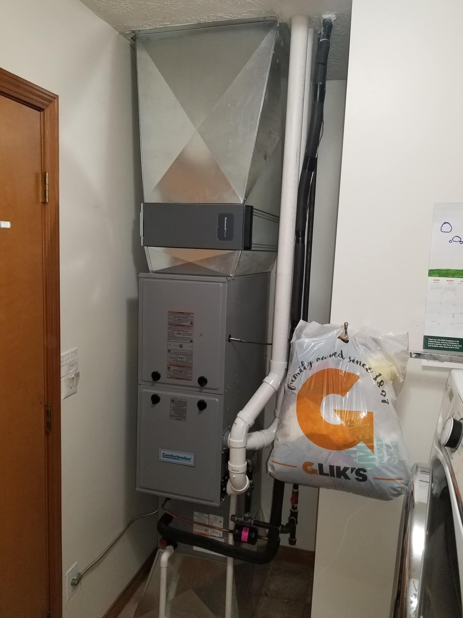 A gray HVAC furnace unit with metal ductwork, white PVC pipes, and a hanging shopping bag in a utility room.