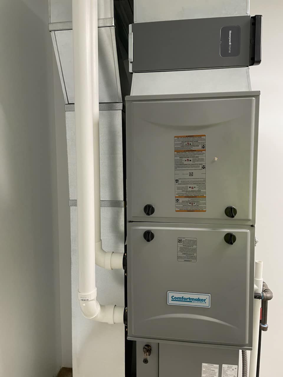 A gray furnace unit with white PVC intake and exhaust pipes installed vertically against a wall.
