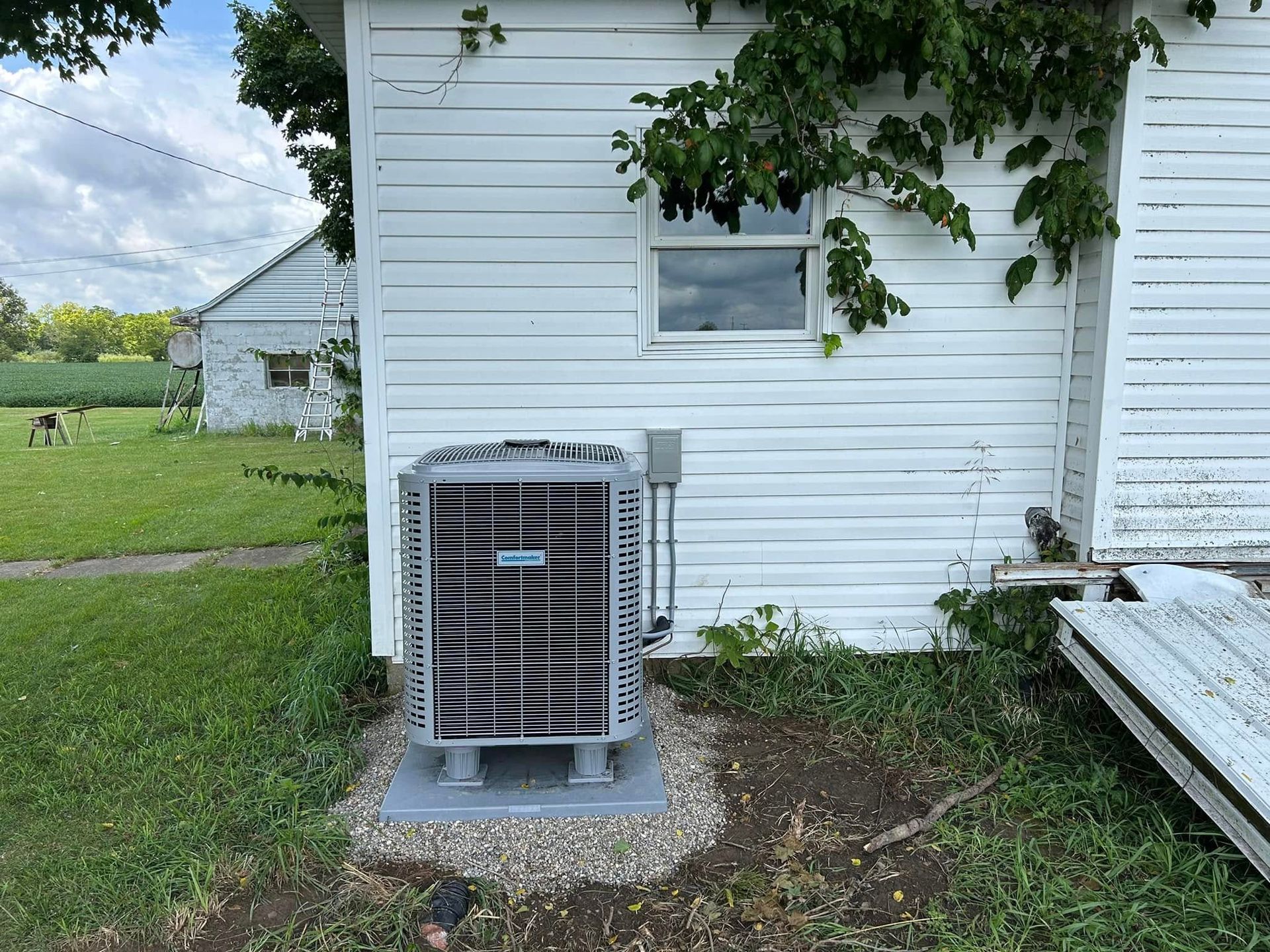A gray HVAC condenser unit sits on a gravel pad next to a white wooden house with green foliage growing on the wall.
