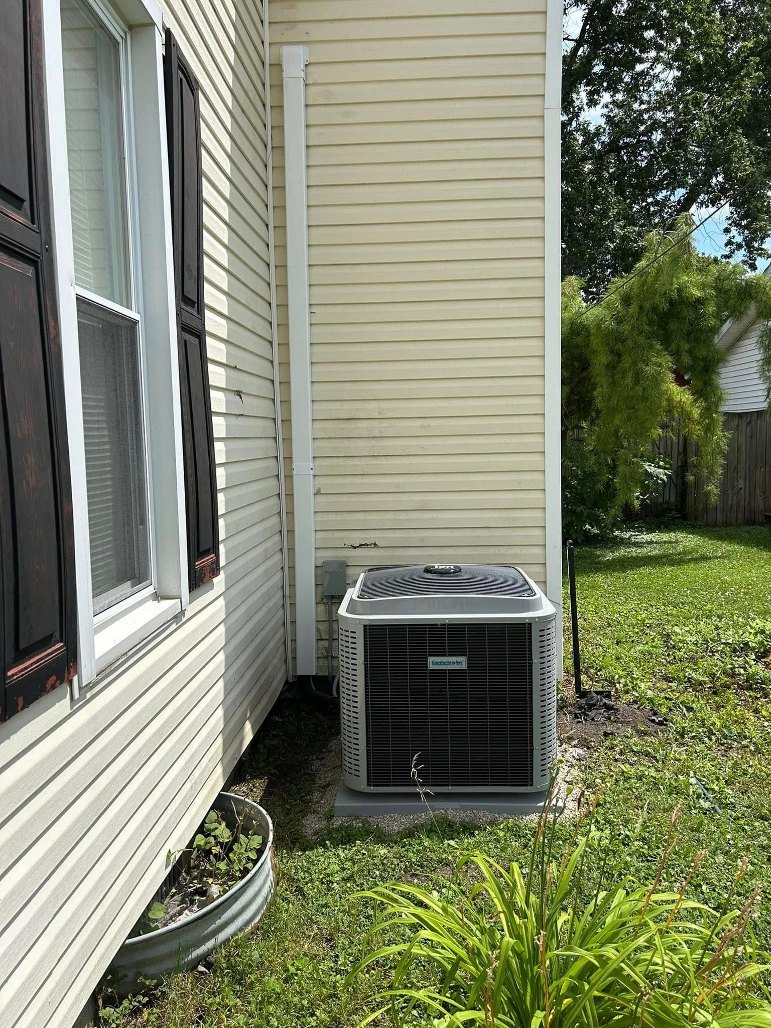 An outdoor HVAC unit sits on a gravel bed next to a yellow-sided house with dark shutters and a white vertical conduit.