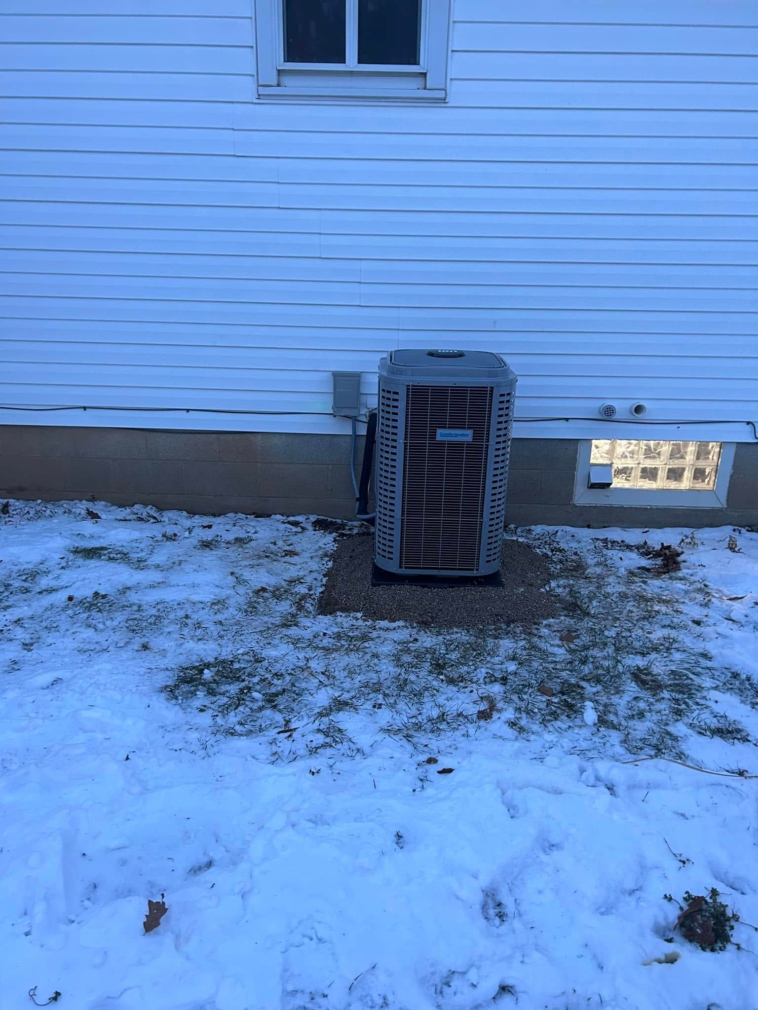 A gray outdoor HVAC unit sits on a gravel pad against a white-sided house covered in snow.
