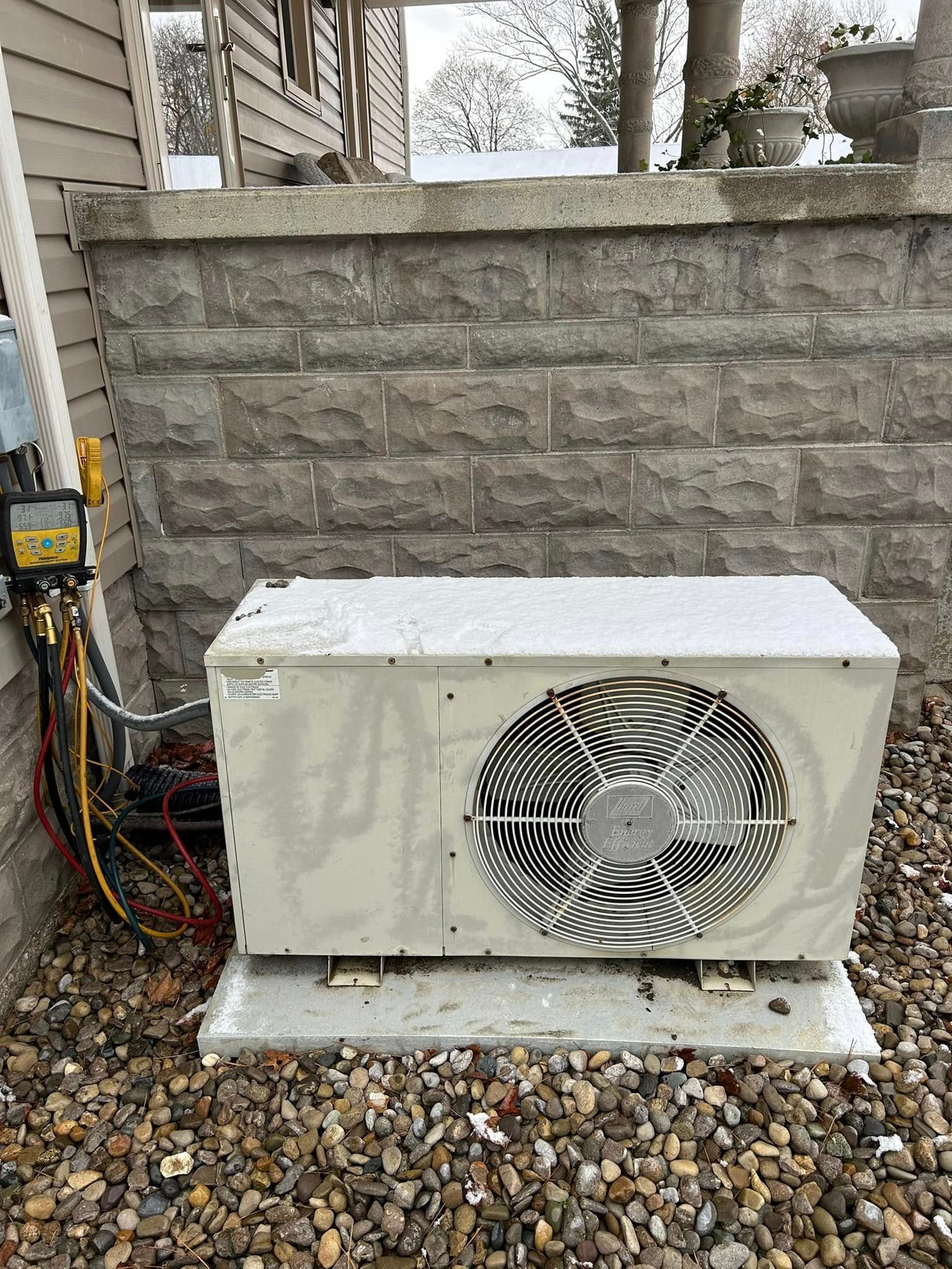 A beige outdoor HVAC unit sits on a concrete pad against a stone wall, connected to diagnostic gauges with colored hoses.