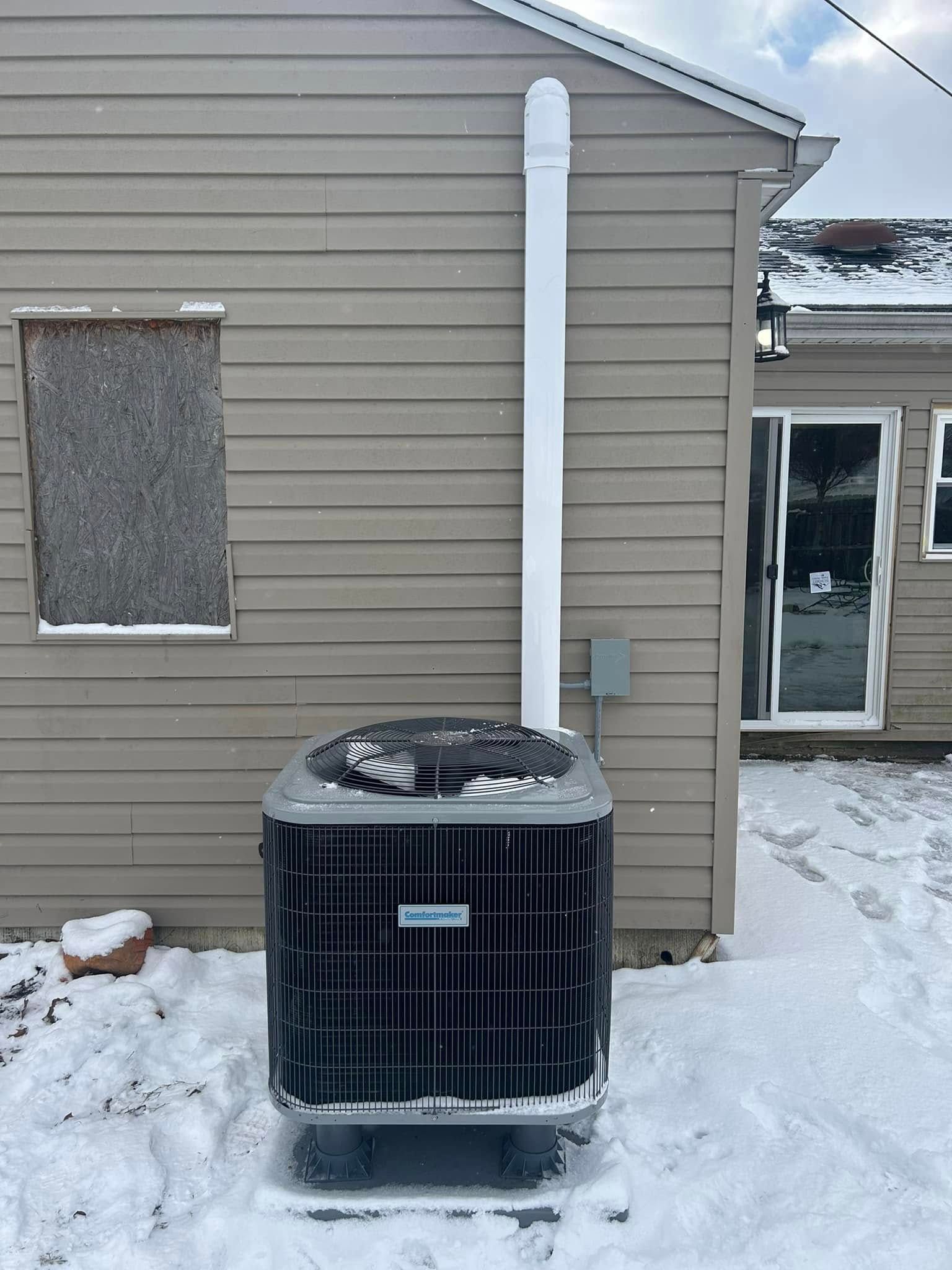 An outdoor HVAC unit sitting in the snow next to a house with a white vertical line set cover on the exterior wall.