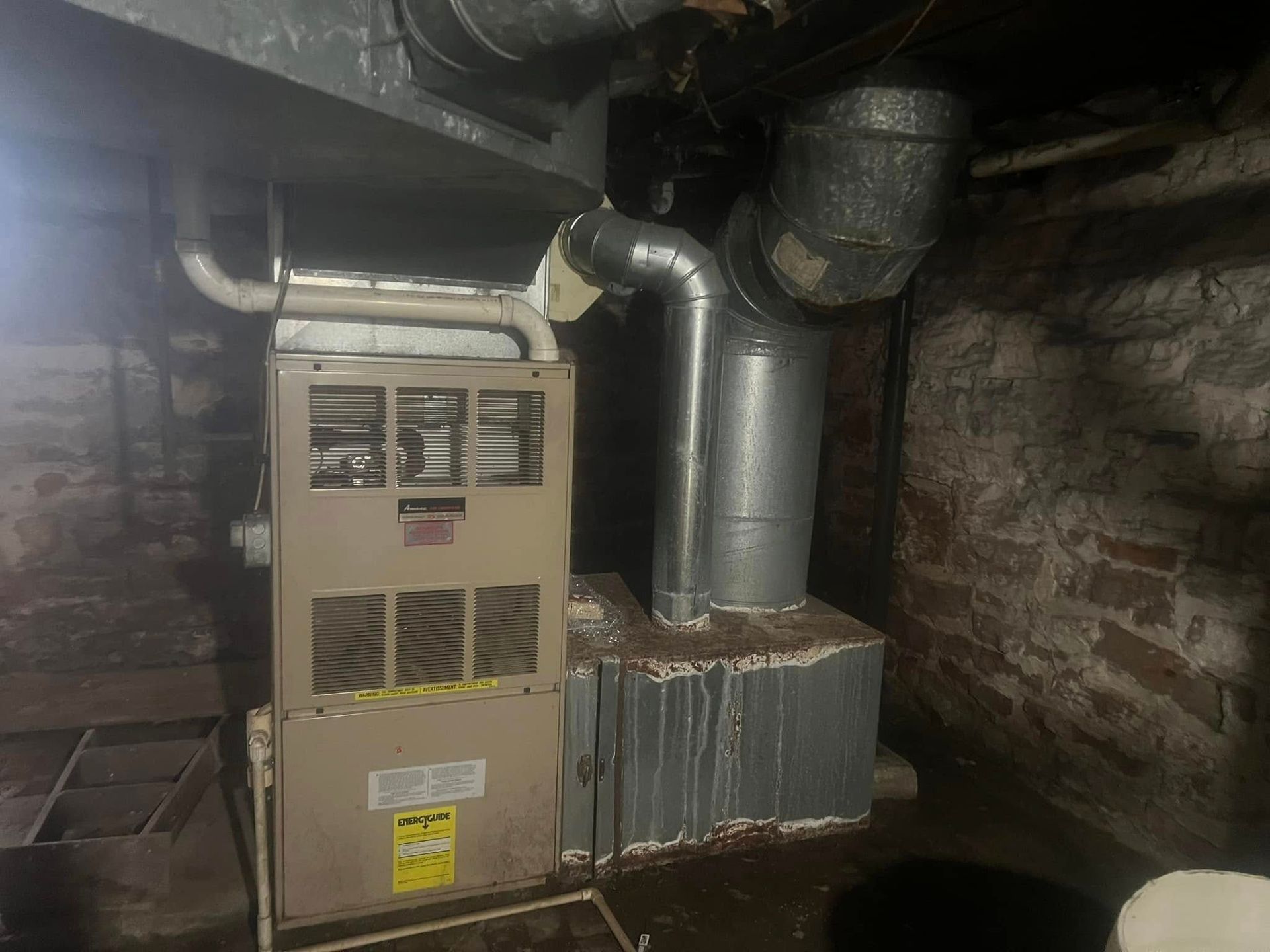 A beige residential furnace in a dimly lit basement with stone walls and silver metal ductwork.