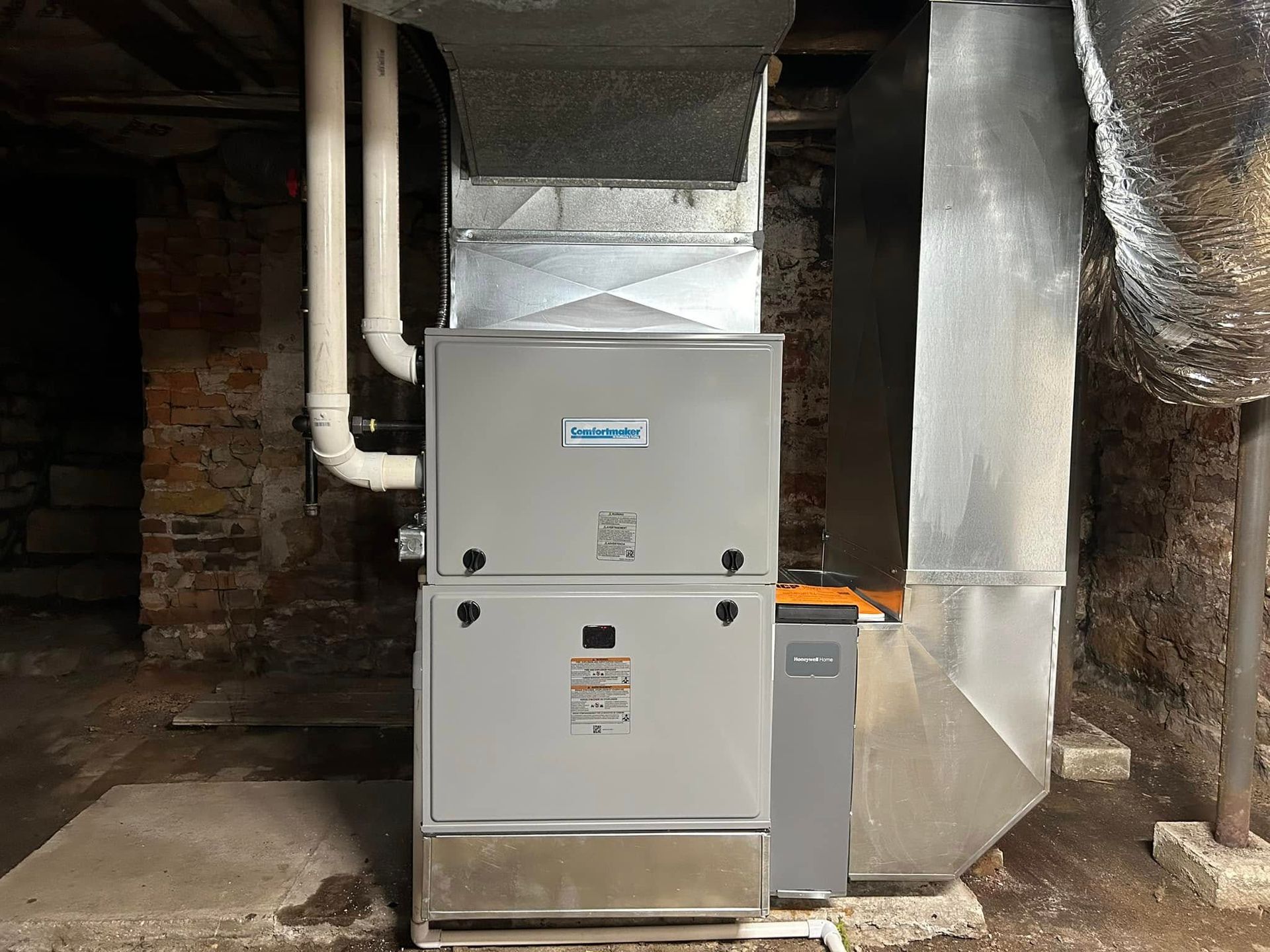 A gray HVAC furnace unit with white vent pipes connected to metal ductwork in a basement with stone walls.