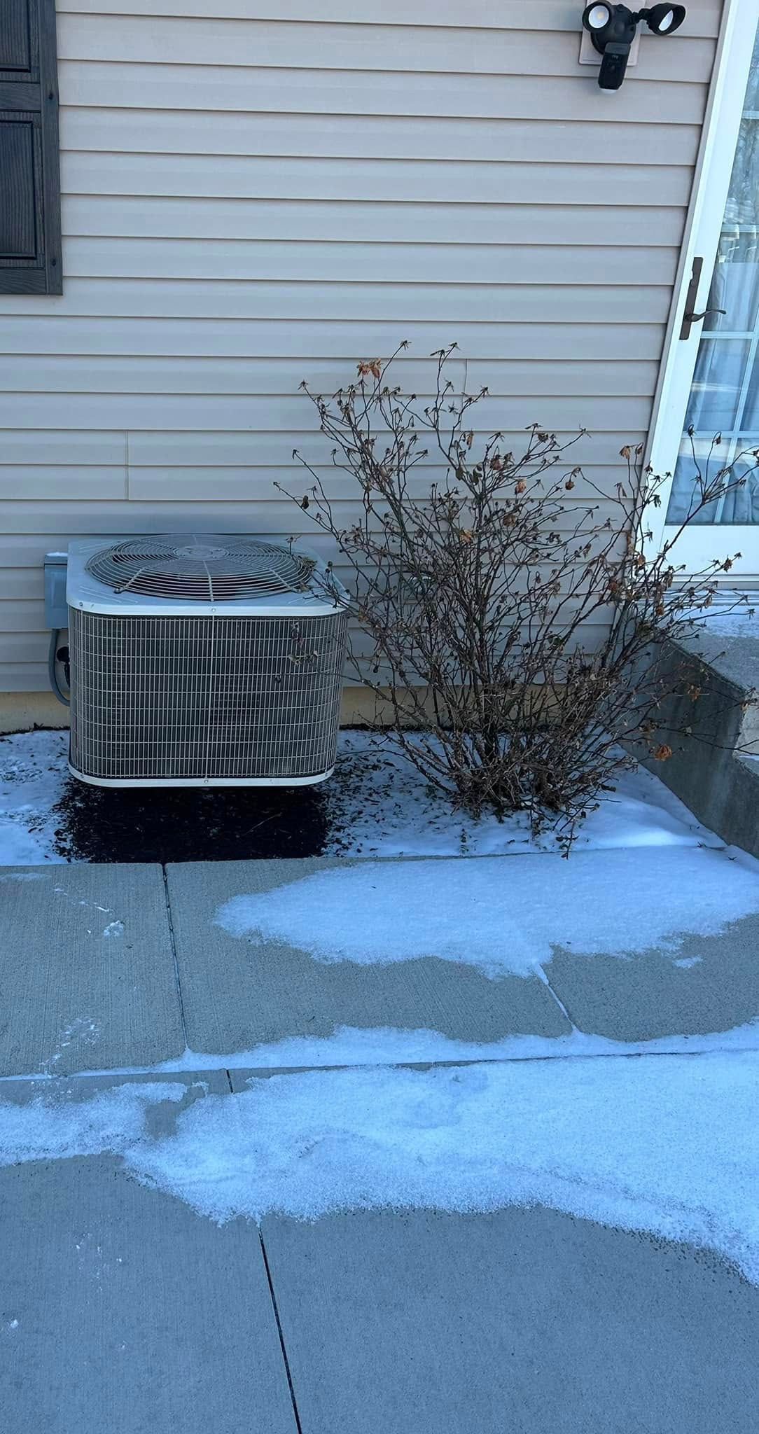 An outdoor air conditioning unit next to a small, dried shrub against a light-sided house, with snow on the ground.