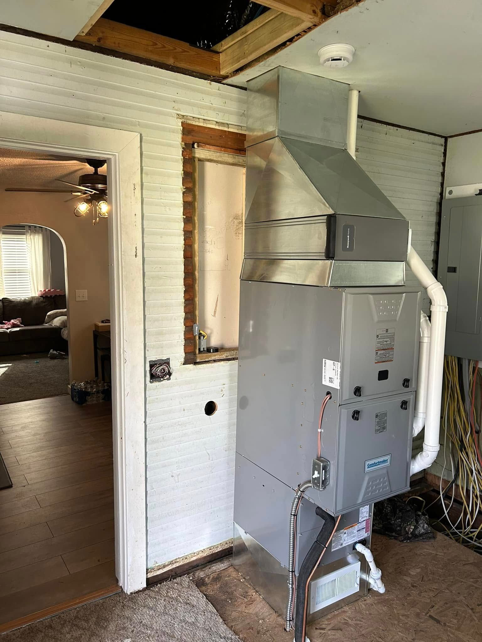 A vertical HVAC furnace unit installed in a room with unfinished walls, near an open doorway and exposed electrical wires.