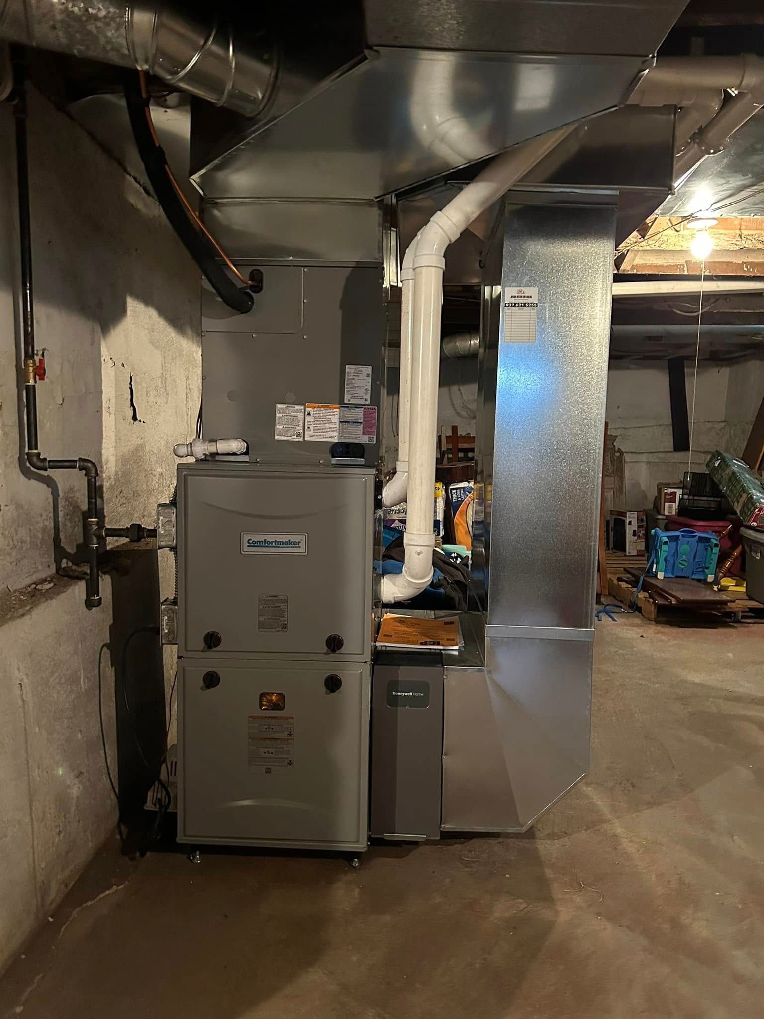 A grey furnace with connected metal ductwork and white PVC exhaust pipes, situated in a basement utility area.