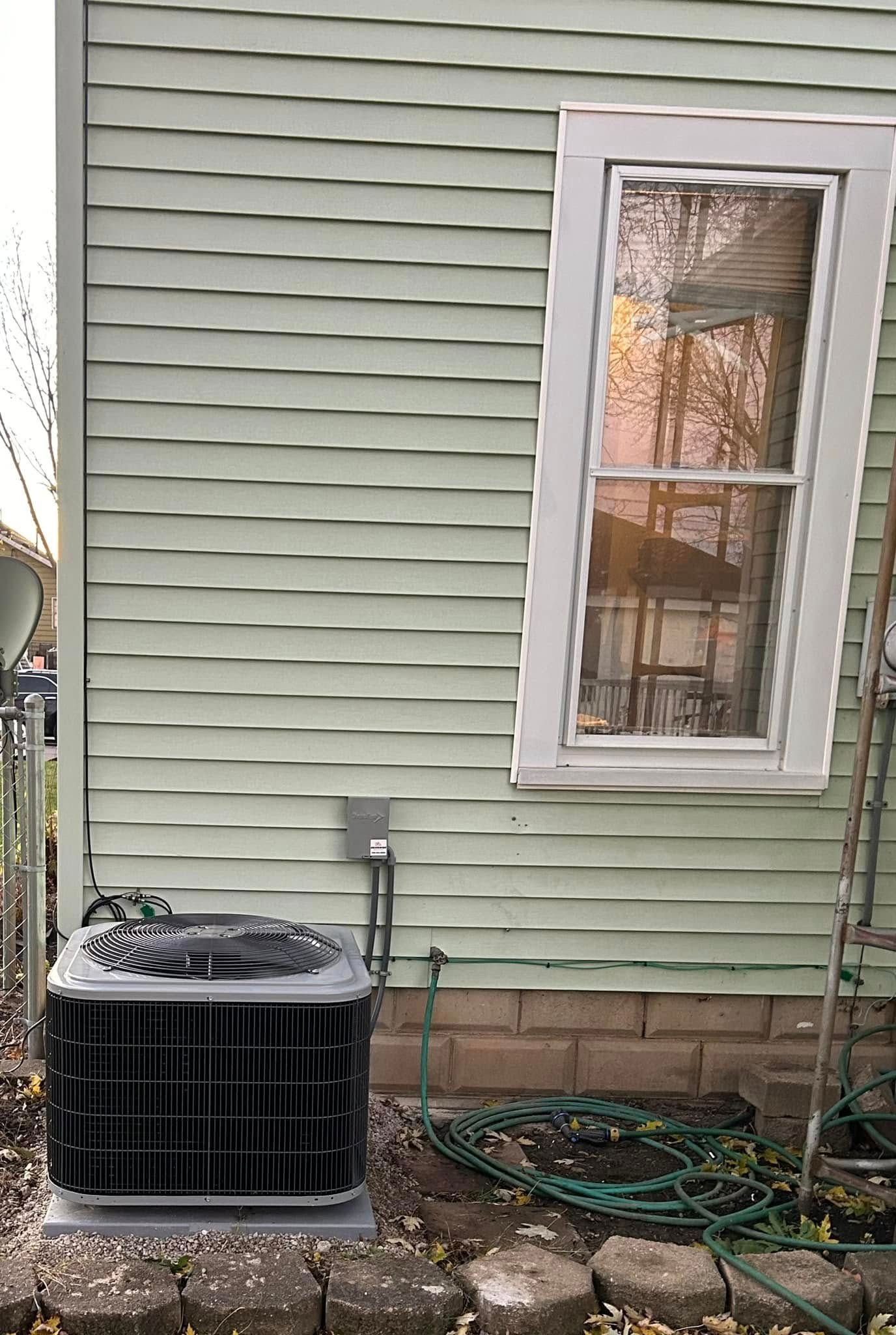 An air conditioning unit sits on the ground outside a house with green siding and a tilted window frame.