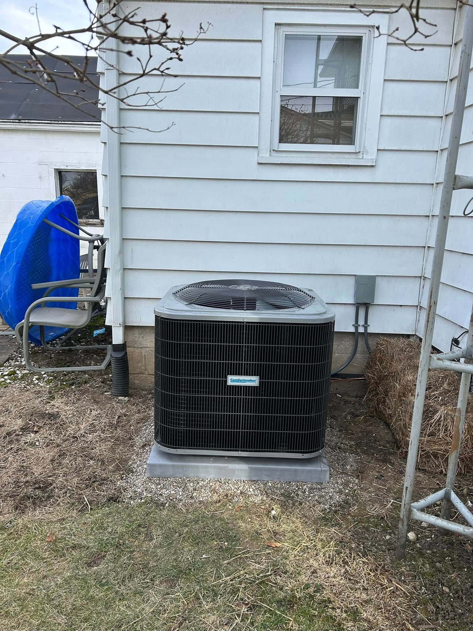 A black outdoor air conditioning unit sits on a concrete pad against a white-sided house with a window above.