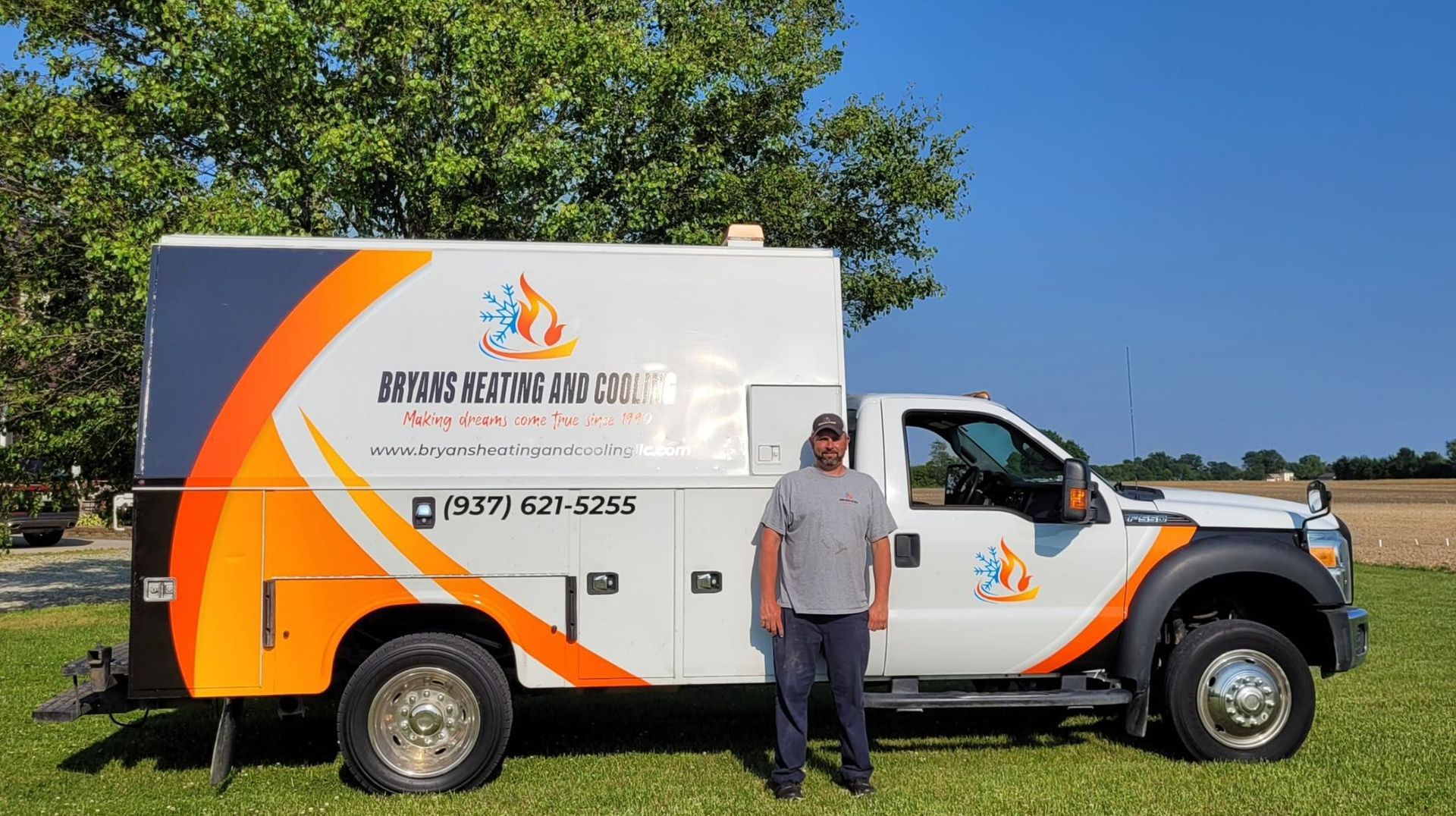 A worker stands beside a white service truck with orange and blue graphics, parked on grass against a blue sky.