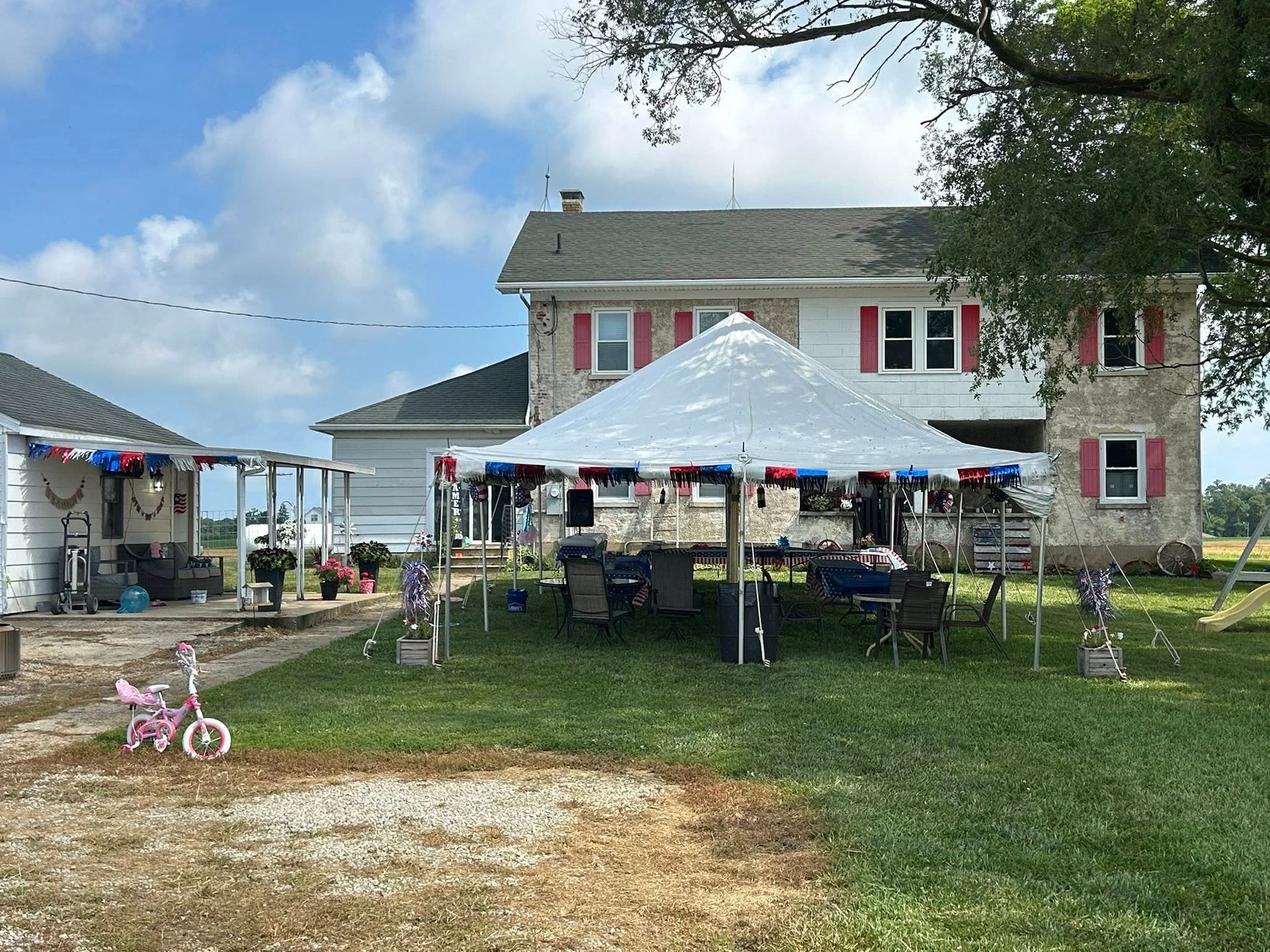 A white party tent decorated with red, white, and blue streamers stands on a grassy lawn in front of a stone farmhouse.