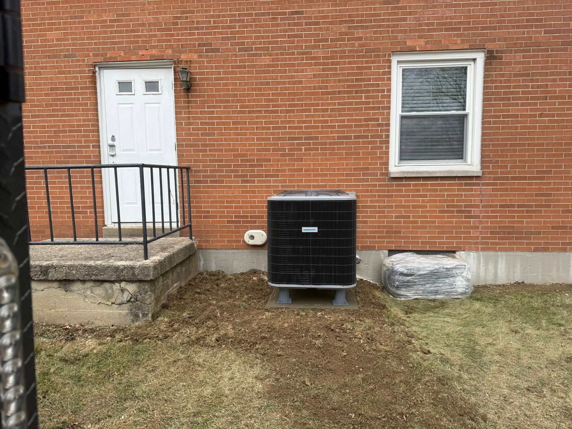 A red brick exterior wall with a white door, a window, and an outdoor HVAC unit sitting on the grass.