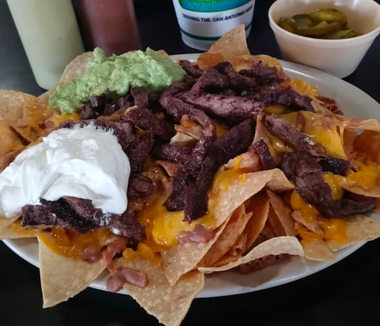 Loaded nachos topped with melted cheese, steak strips, sour cream, guacamole, and jalapeños on a plate.